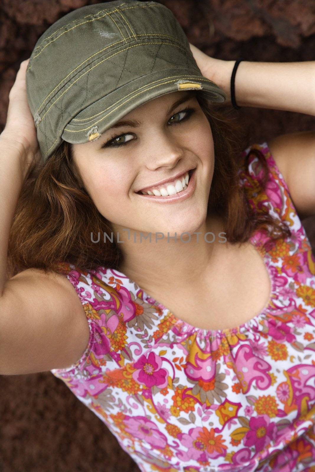 Young Caucasian woman wearing hat with hands behind head smiling at viewer.