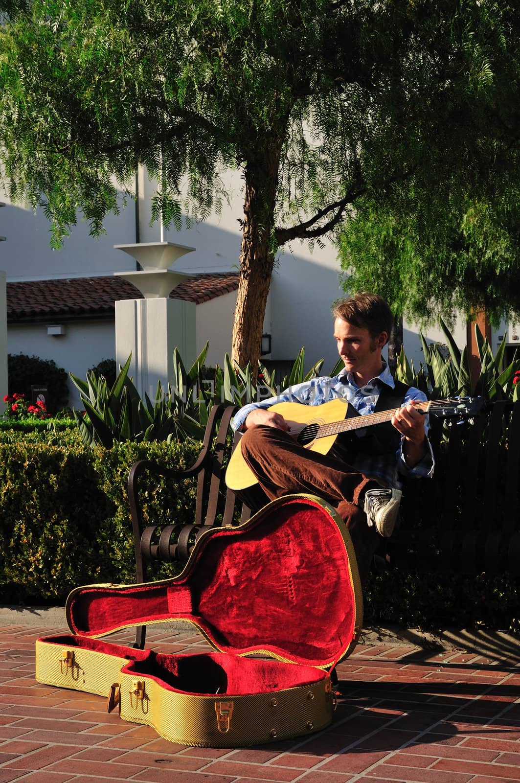 Young man playing music on his guitar for donations in a sunny courtyard.