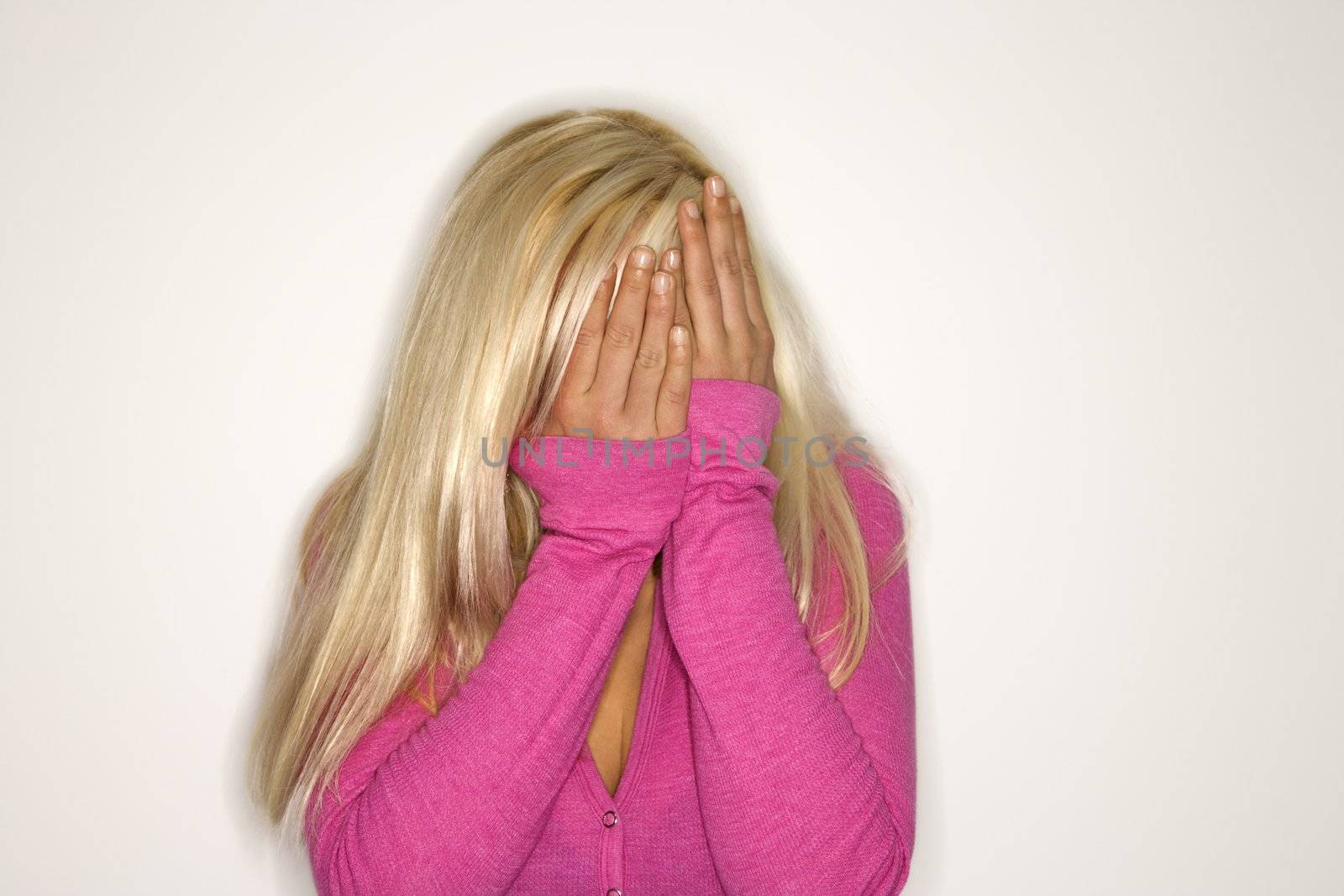 Portrait of attractive blonde Caucasian young adult woman wearing fuchsia covering face with hands.