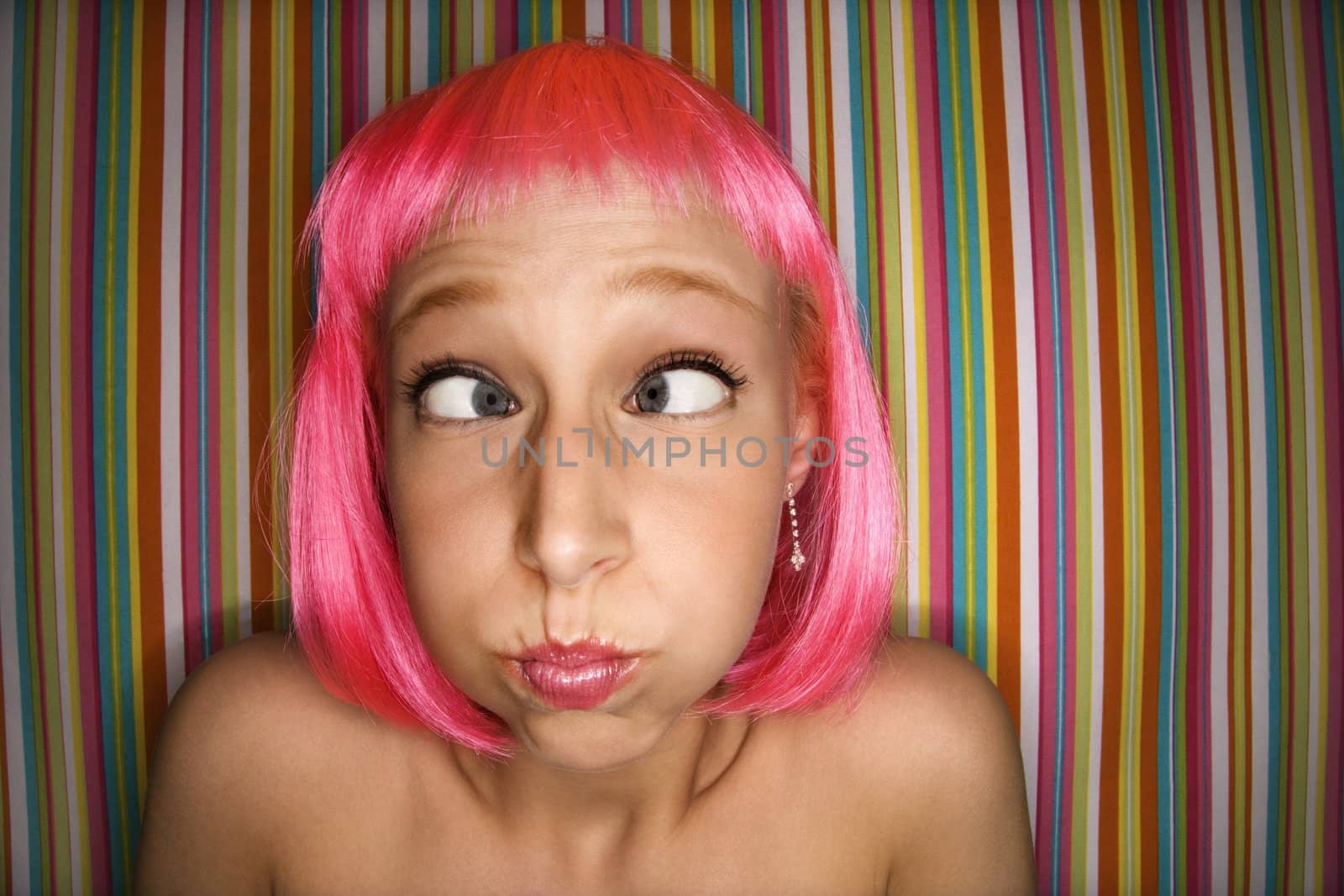 Portrait of attractive Caucasian young adult woman wearing pink wig against striped background making cross eyed expression.
