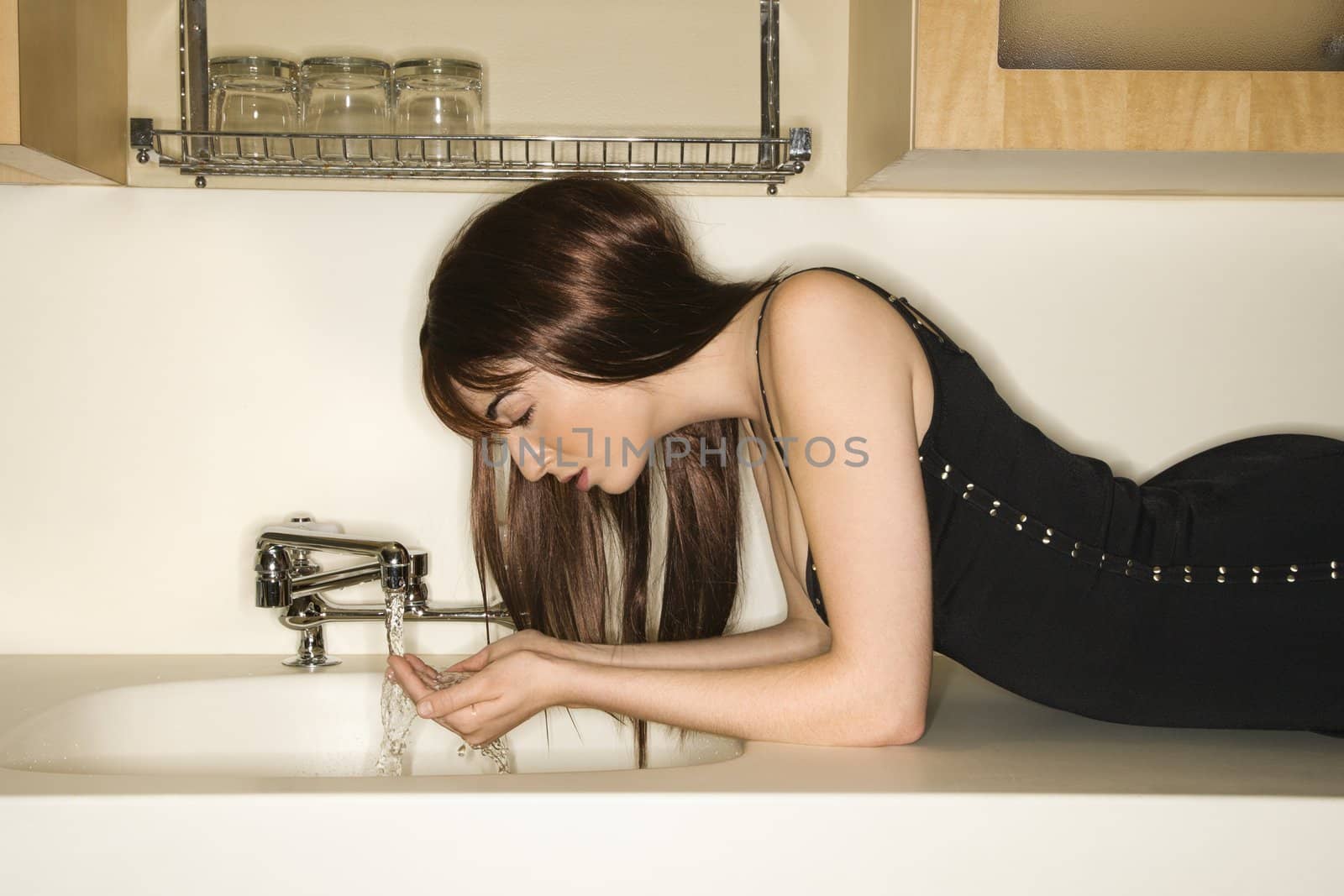 Pretty Caucasian young woman lying on kitchen counter drinking water from faucet.