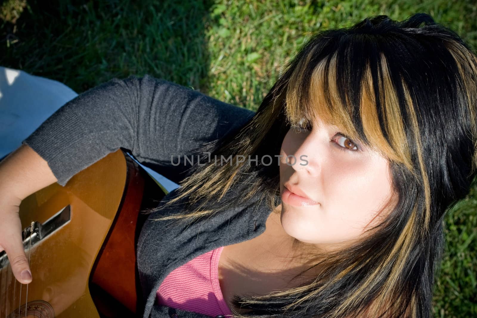 A young hispanic woman playing a guitar while laying on a blanket in the green grass.  Her hair is highlighted with blonde streaks.