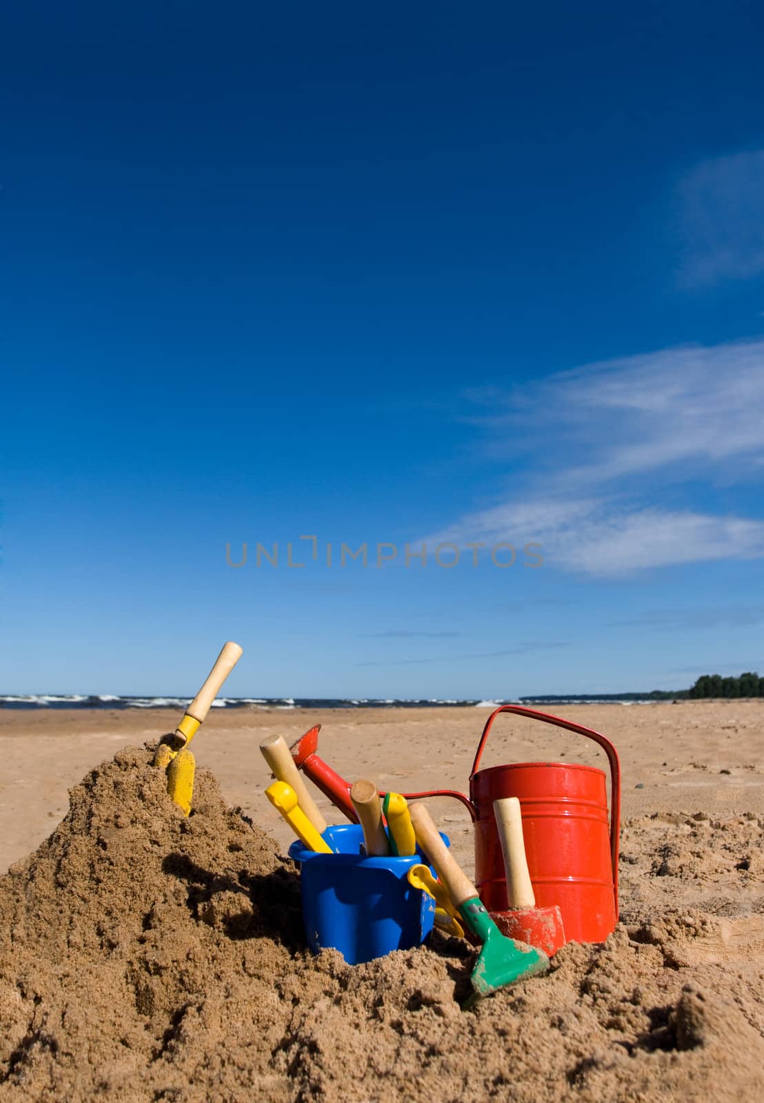 Red watering can, plastic blue bucket and other beach toys in the sandy seashore