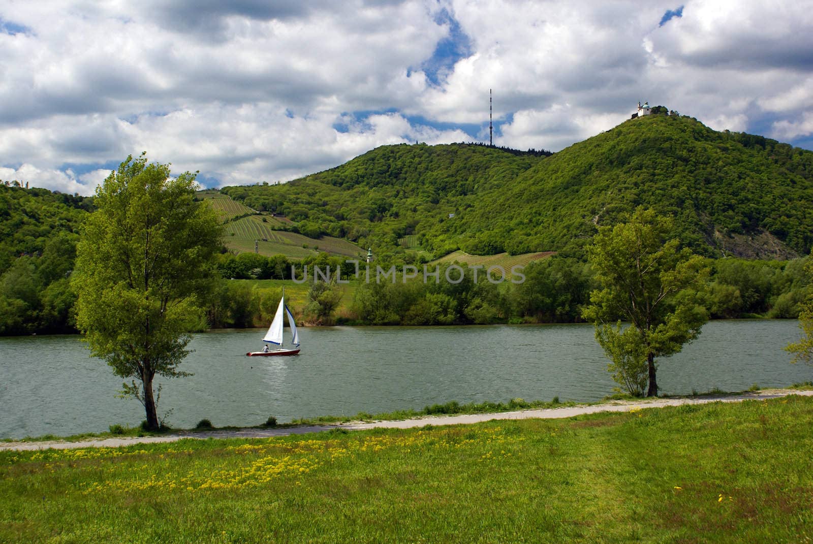 A little sail boat on a river green hills in the background cloudy sky.