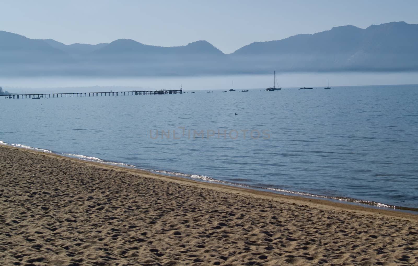 Lake shore with boats and pier in the background, mountains and fog in the distance.