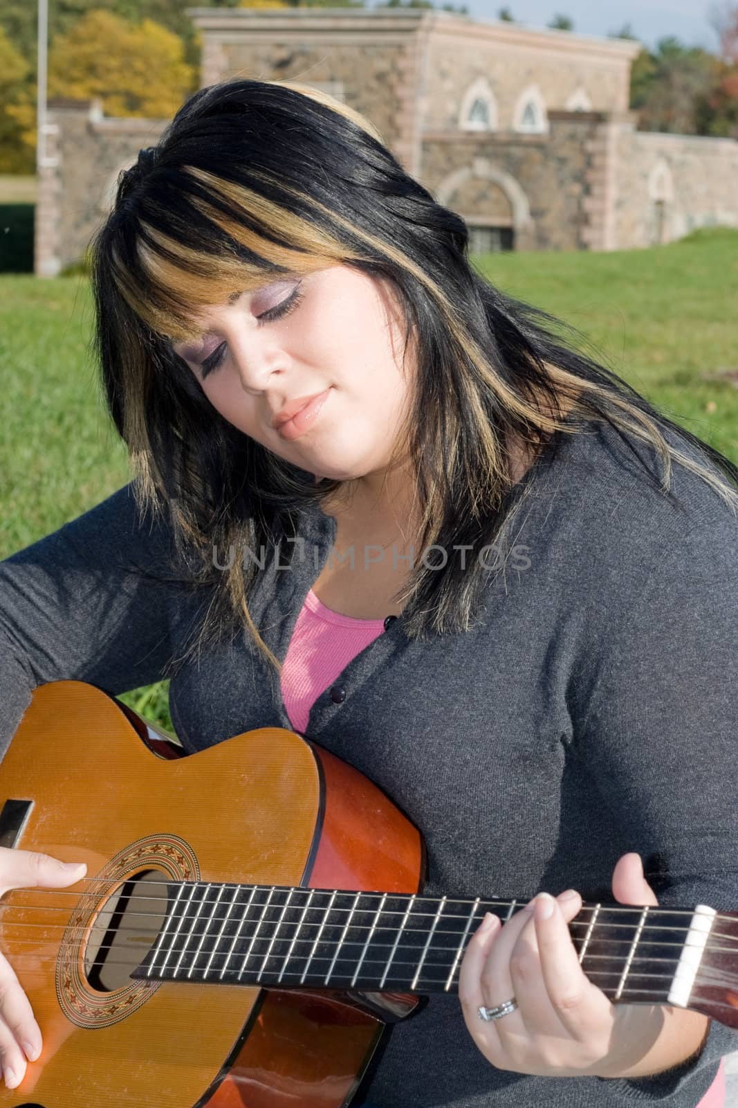A young hispanic woman playing a guitar while sitting on a blanket in the green grass. Her hair is highlighted with blonde streaks.