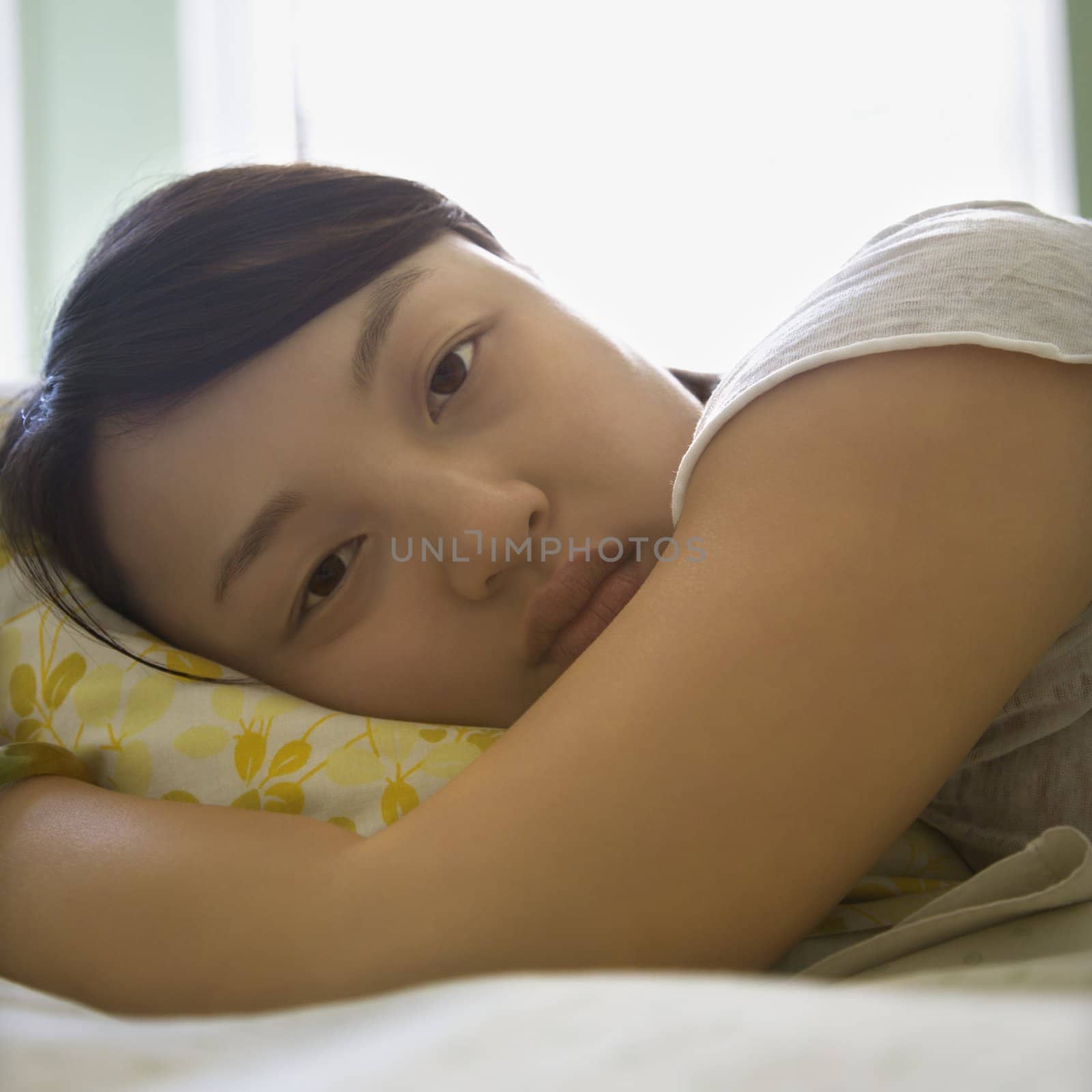 Portrait of pretty young Asian woman lying in bed with head on pillow making eye contact.