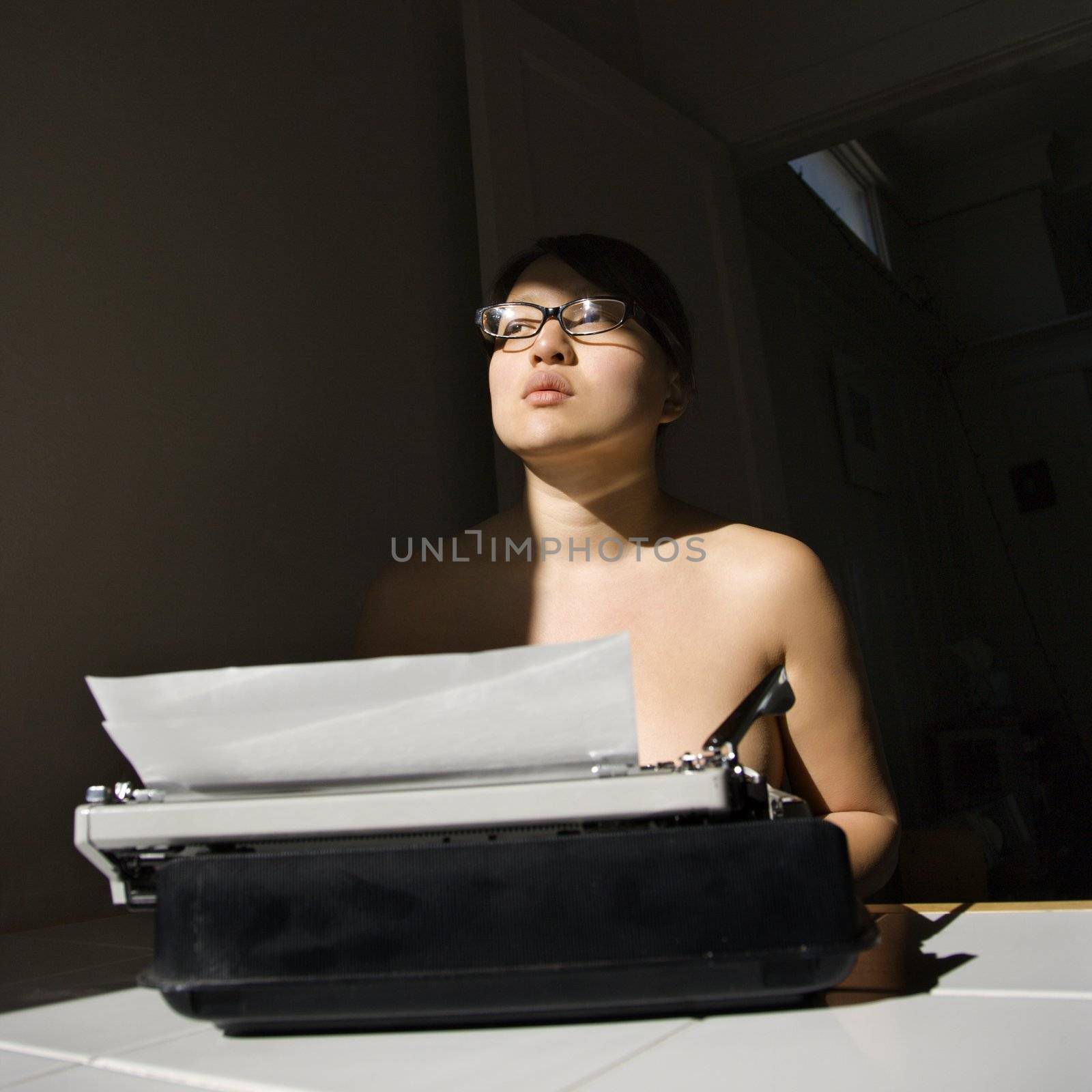 Nude young Asian woman sitting at kitchen table with typewriter.