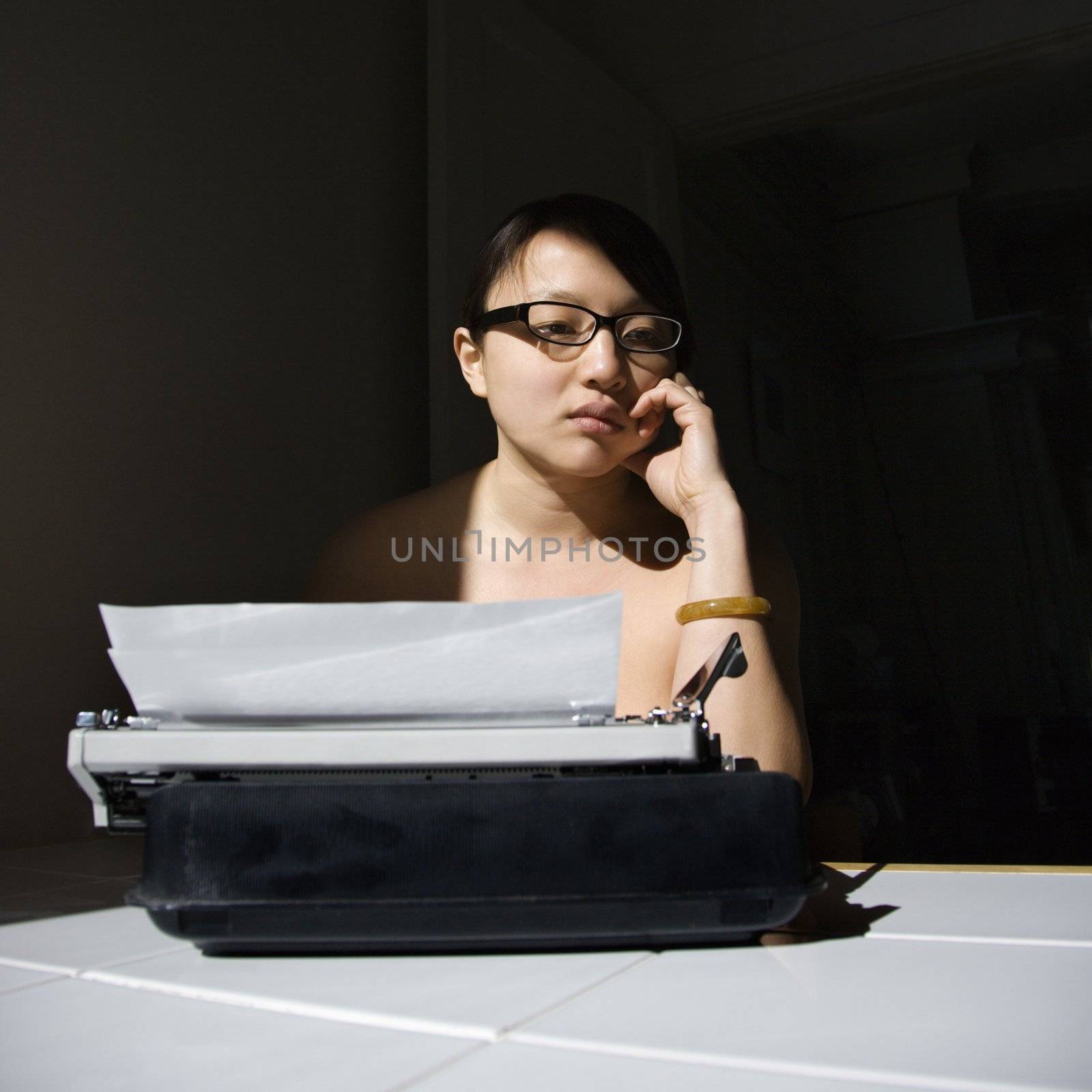 Nude young Asian woman sitting at kitchen table with typewriter.
