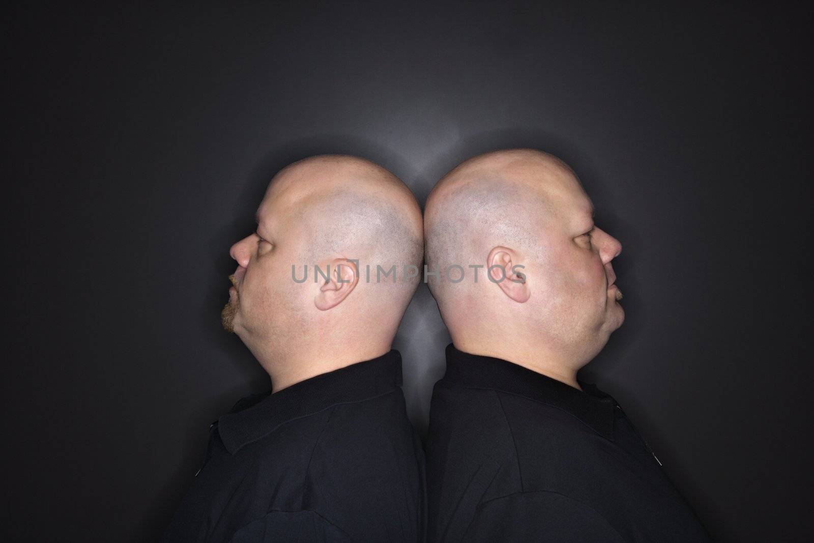 Caucasian mid adult identical twin bald men standing back to back.