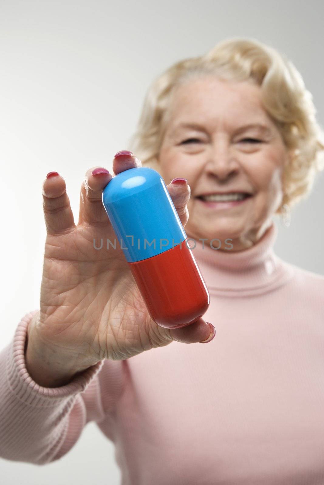 Caucasian senior woman holding oversized pill at viewer.