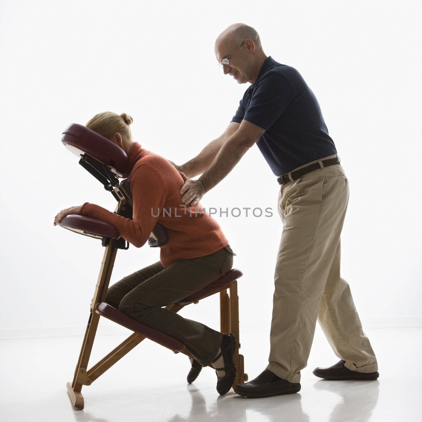 Caucasian middle-aged male massage therapist massaging back of Caucasian middle-aged woman sitting in massage chair.