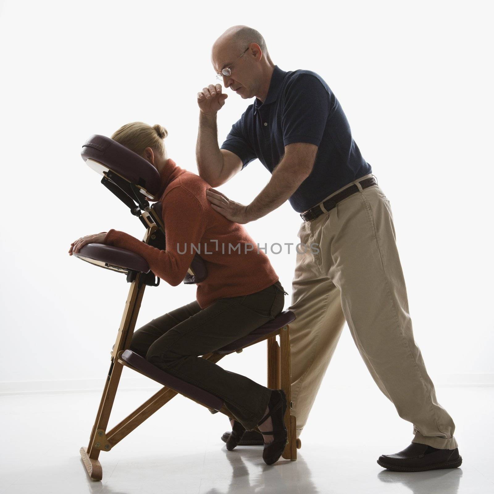 Caucasian middle-aged male massage therapist massaging back of Caucasian middle-aged woman sitting in massage chair with his elbow.