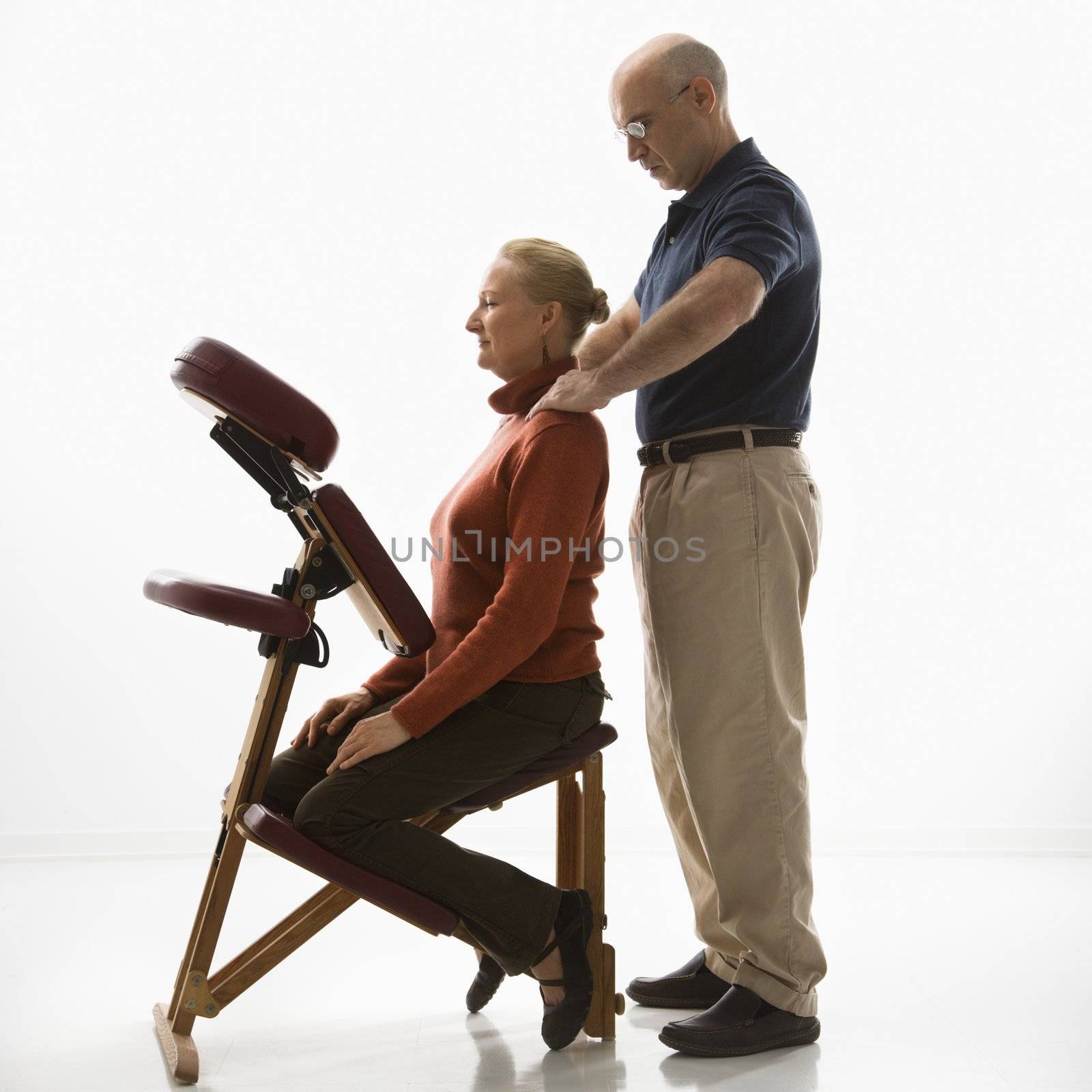 Caucasian middle-aged male massage therapist massaging shoulders of Caucasian middle-aged woman sitting in massage chair.