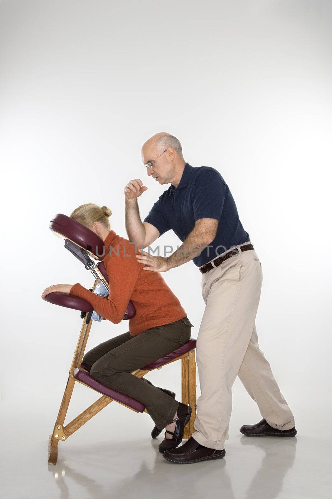 Caucasian middle-aged male massage therapist massaging back of Caucasian middle-aged woman sitting in massage chair with his elbow.