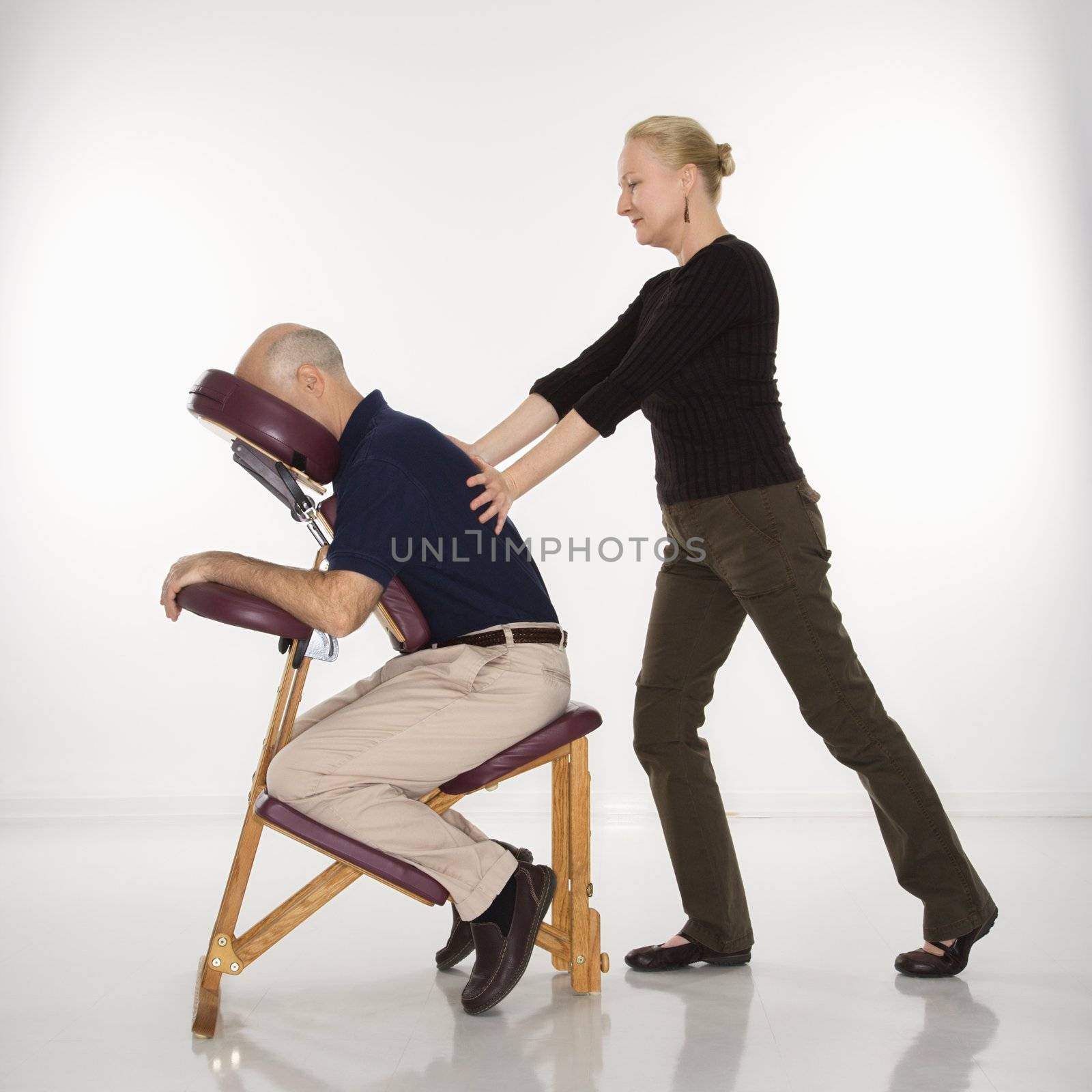Caucasian middle-aged female massage therapist massaging back of Caucasian middle-aged man sitting in massage chair.