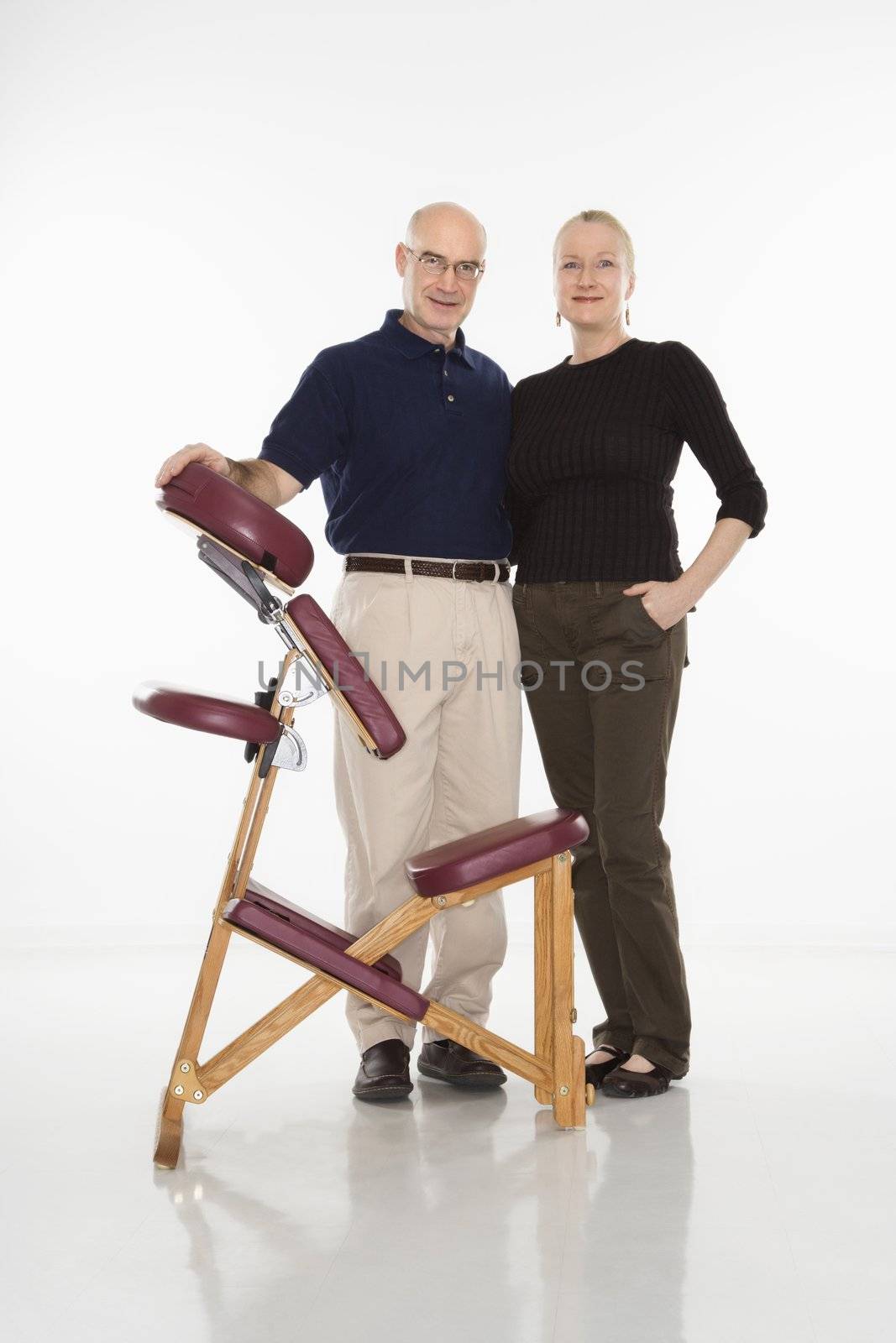 Caucasian middle-aged male massage therapist standing with arm around Caucasian middle-aged woman beside massage chair.