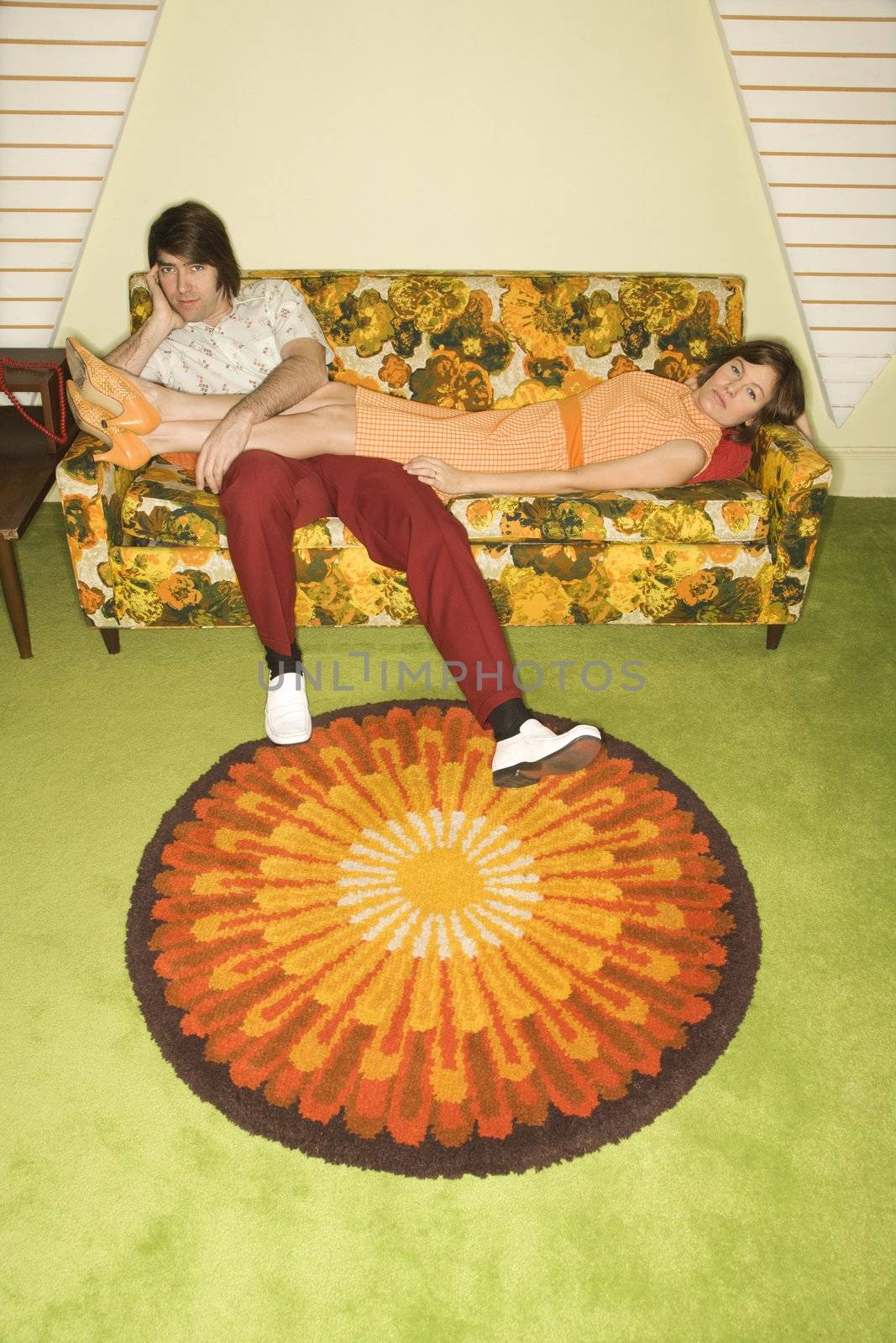 Caucasian mid-adult woman lying on colorful retro sofa with legs draped over Caucasian mid-adult man.