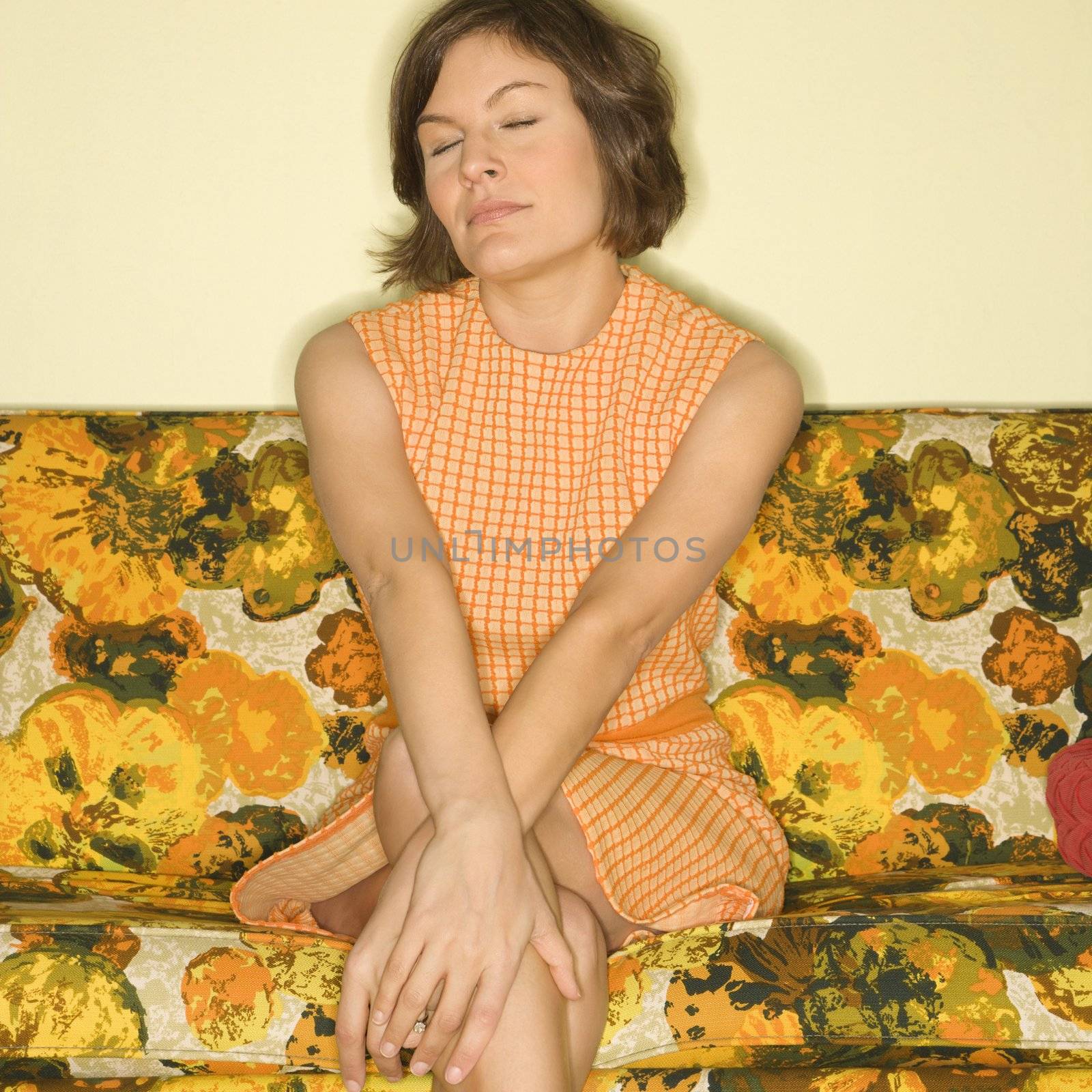 Pretty Caucasian mid-adult woman sitting on colorful retro sofa with arms crossed and eyes closed.