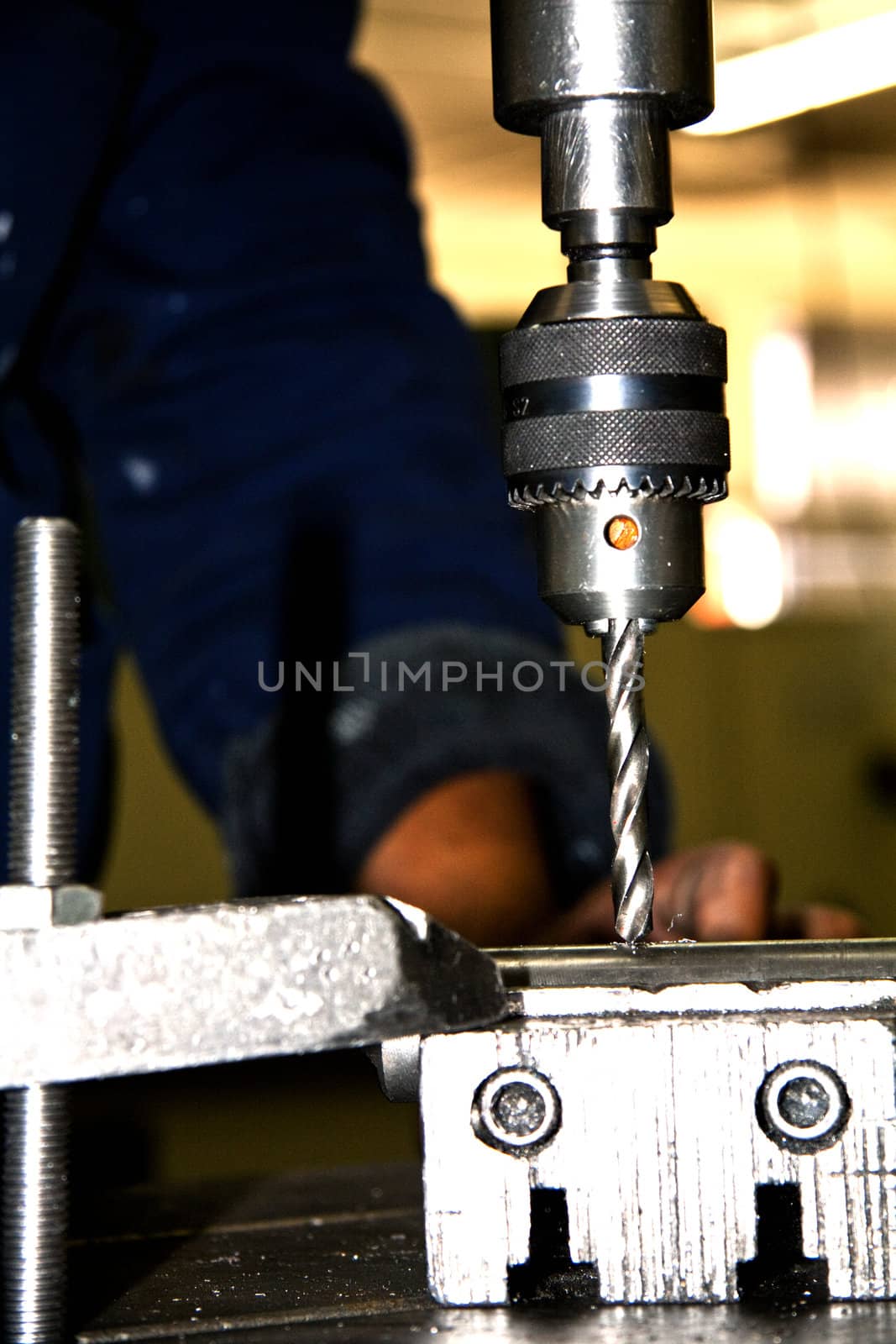 Drill press boring through a piece of metal, showing a man's hand in the background