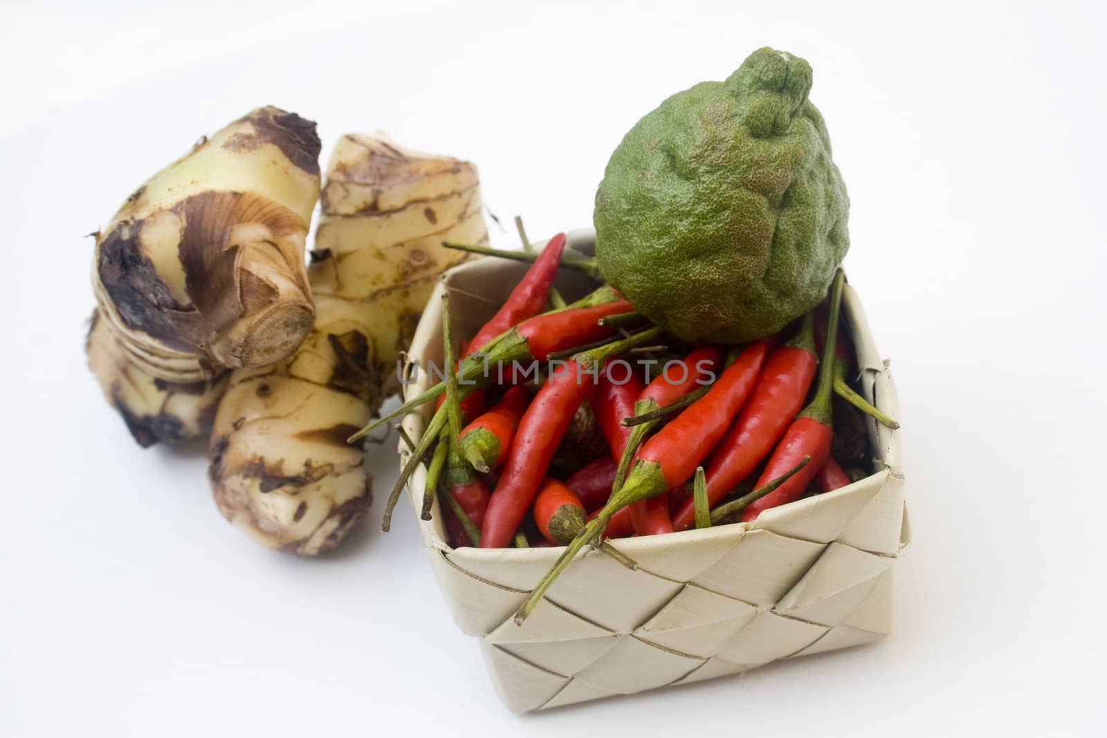 Basket of small chili peppers, galangal root, and a kaffir lime isolated against white