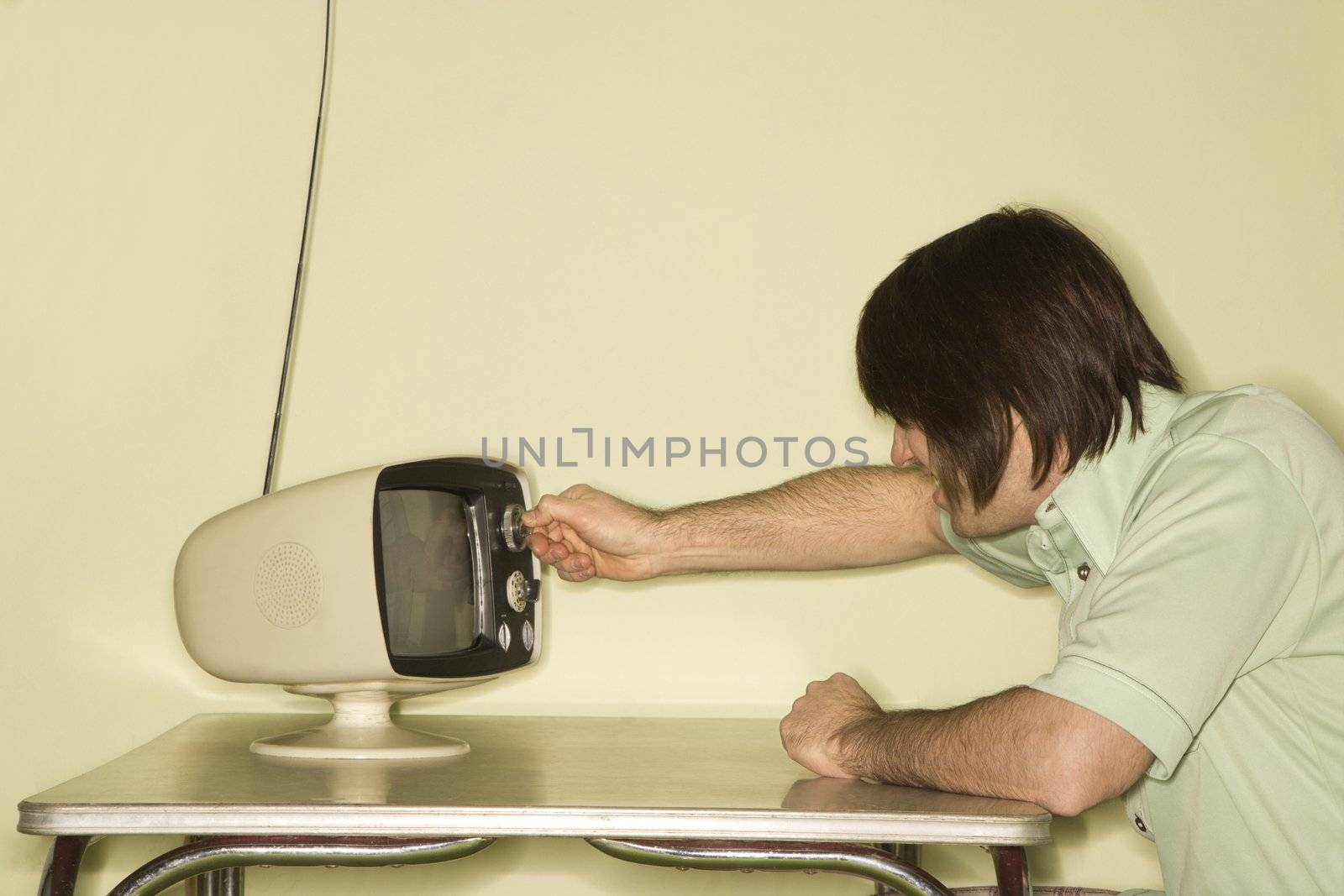 Side view of Caucasian mid-adult man sitting at 50's retro dinette set turning old television knob.