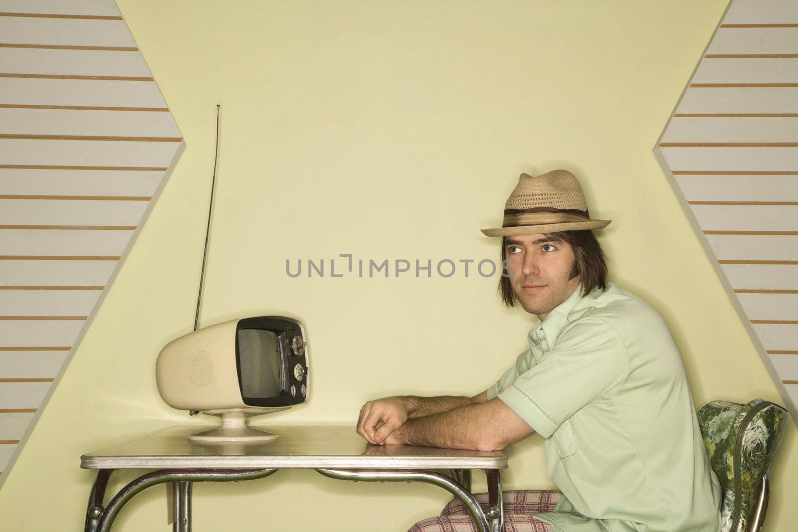 Caucasian mid-adult man wearing hat sitting at 50's retro dinette set in front of old television.