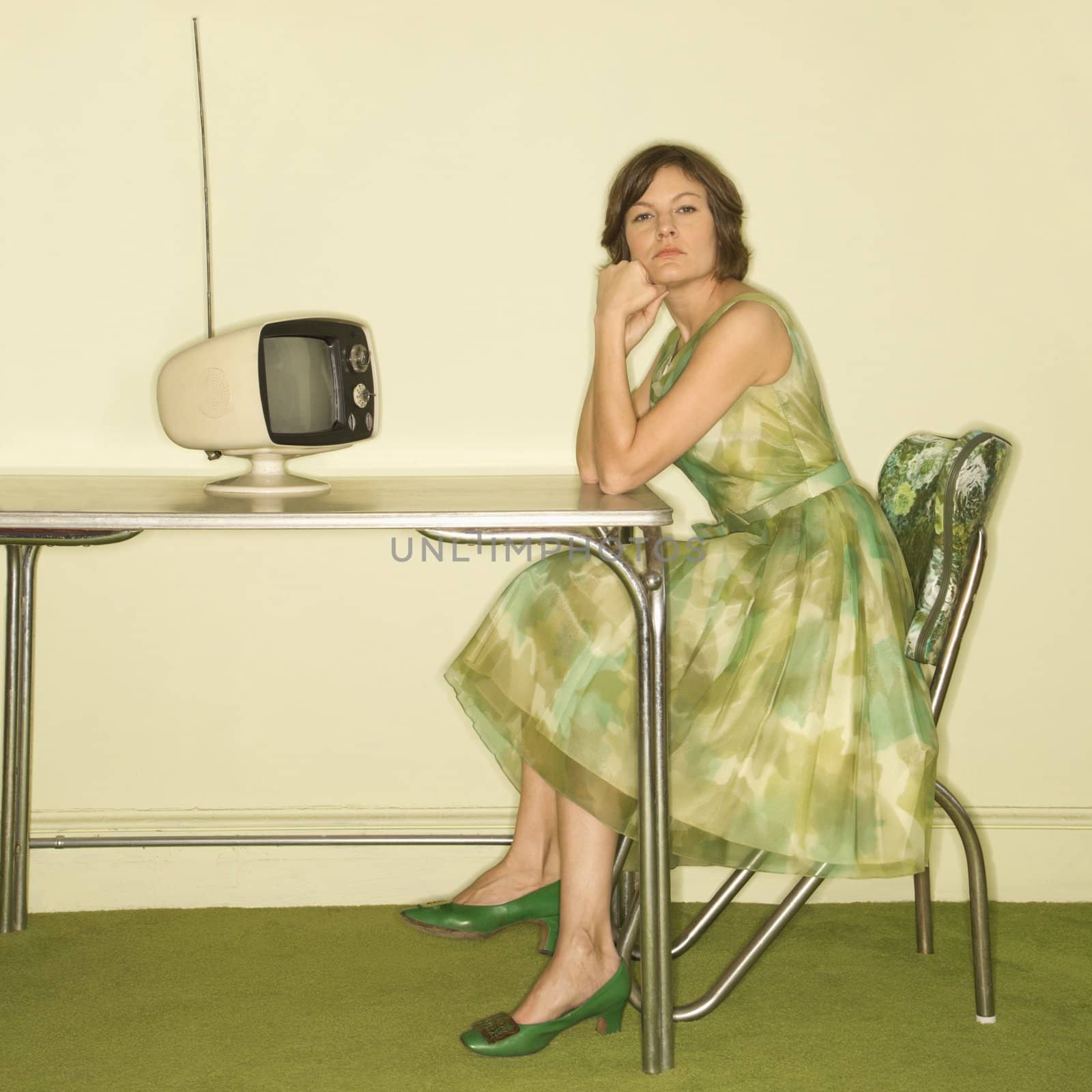 Pretty Caucasian mid-adult woman wearing green vintage dress sitting at 50's retro dinette in front of old televsion looking bored.