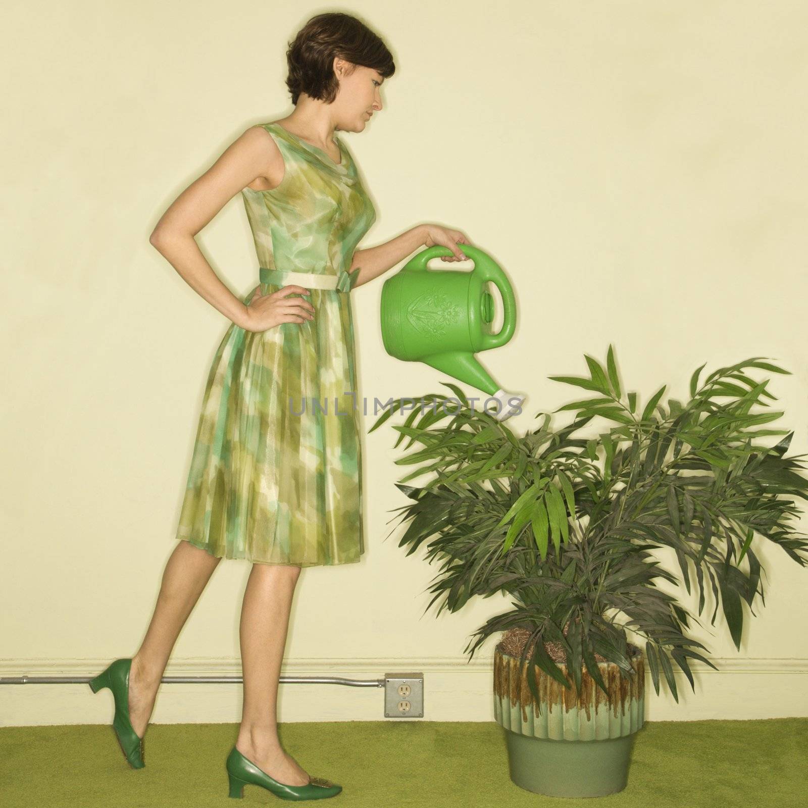 Pretty Caucasian mid-adult woman wearing vintage dress watering houseplant with green watering can.