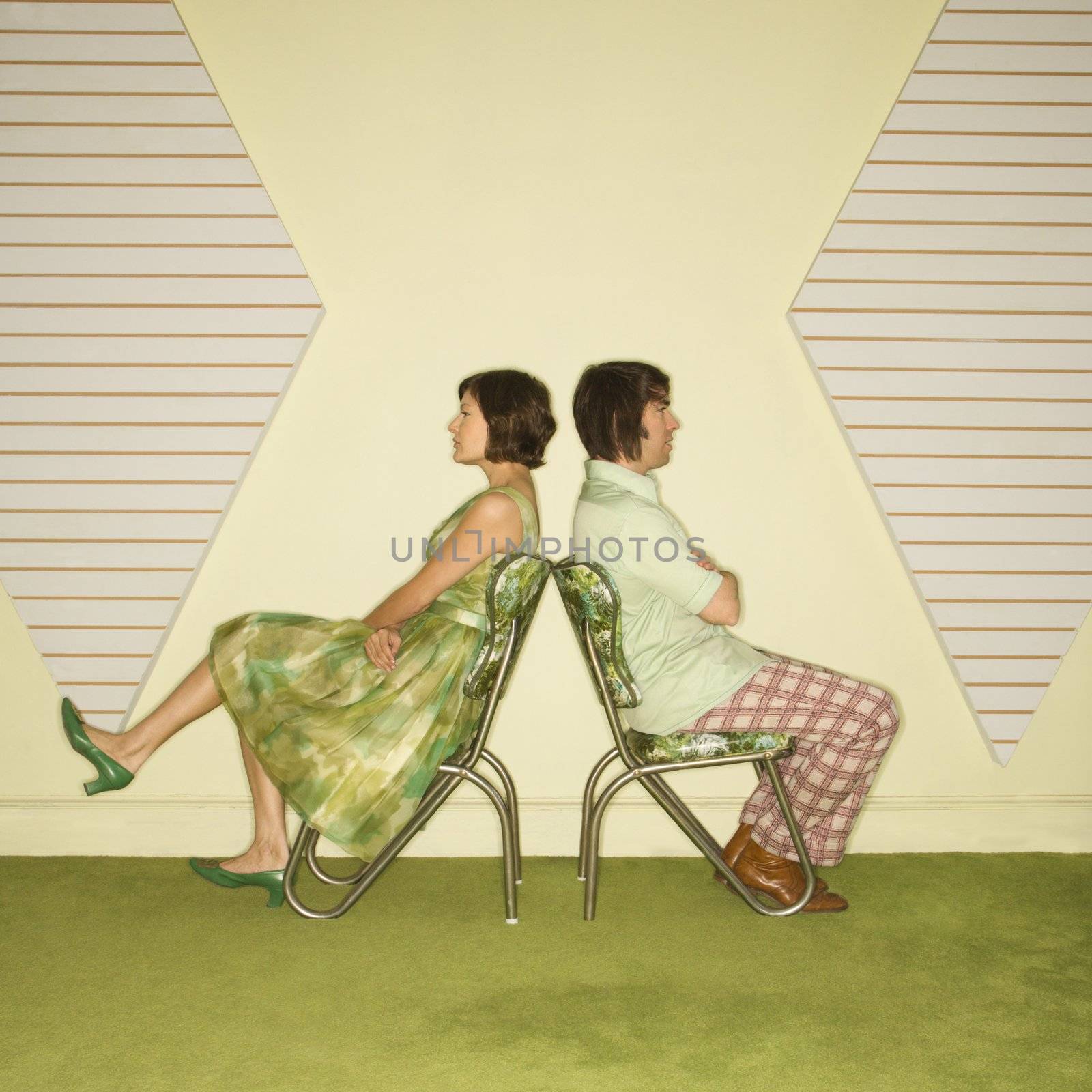 Caucasian mid-adult couple wearing vintage clothing sitting back to back in green vinyl chairs with arms crossed.