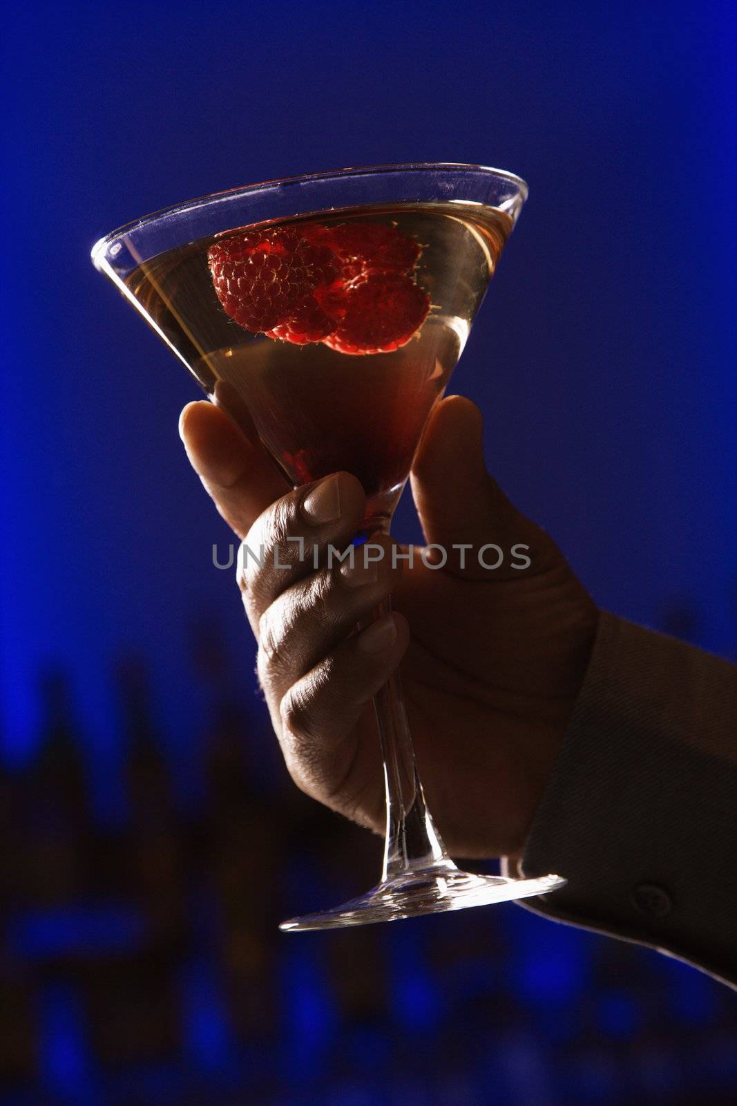 African American man's hand holding holding martini mixed drink with raspberry fruit against glowing blue background.