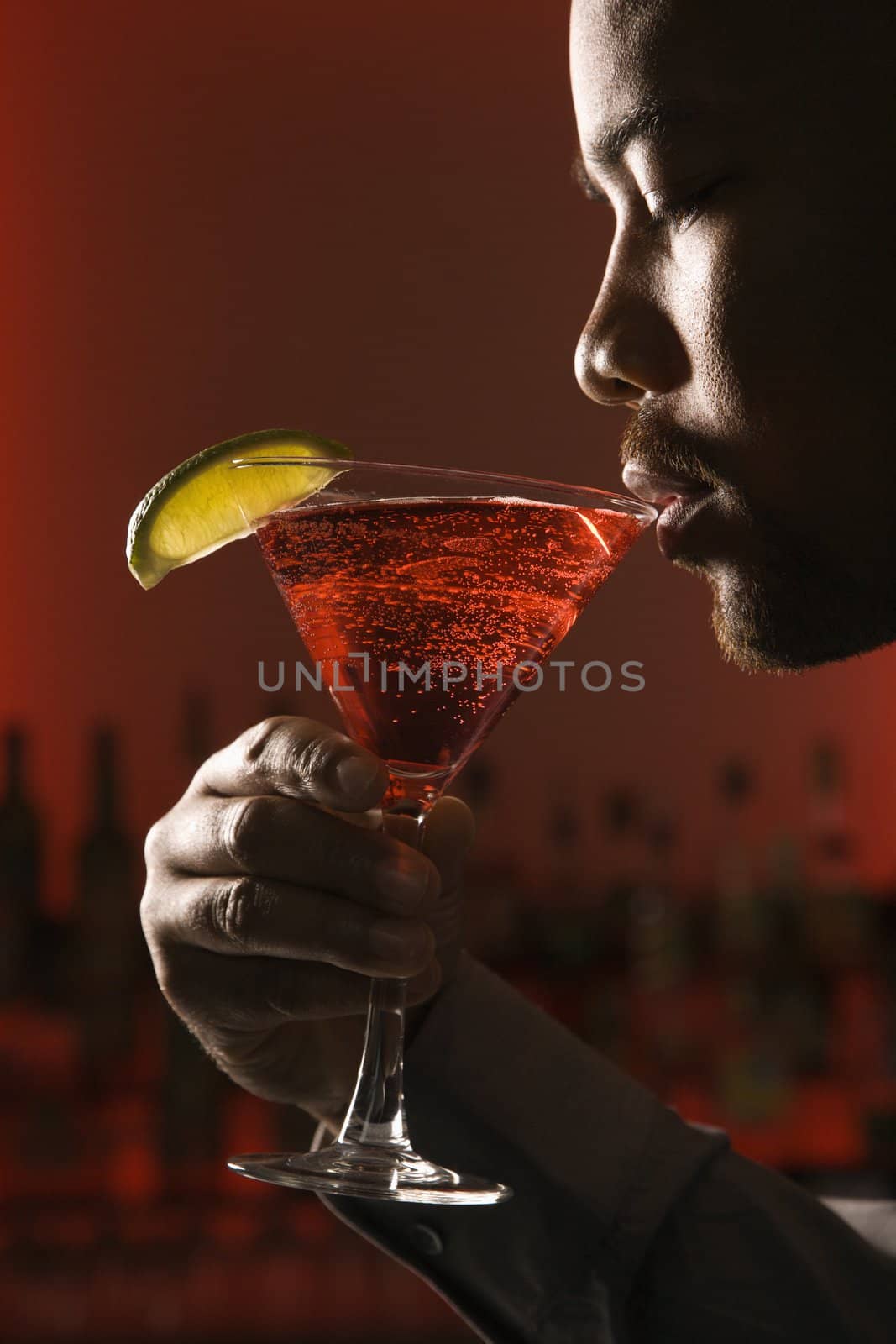 African American man drinking martini in bar against glowing red background.