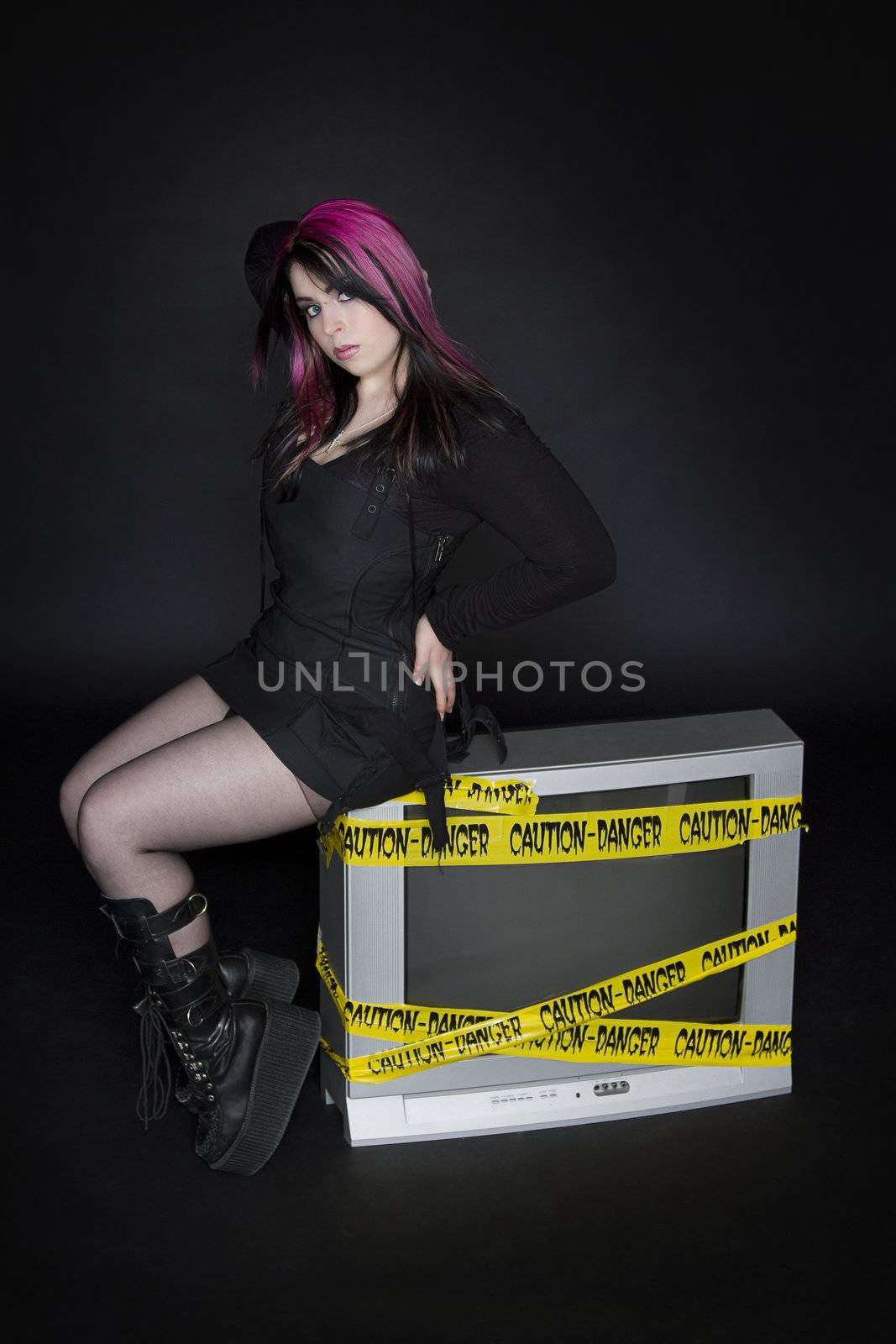 teenage girl wearing goth inspired clothes with pink and black hair in sitting on a tv taking a pin up pose