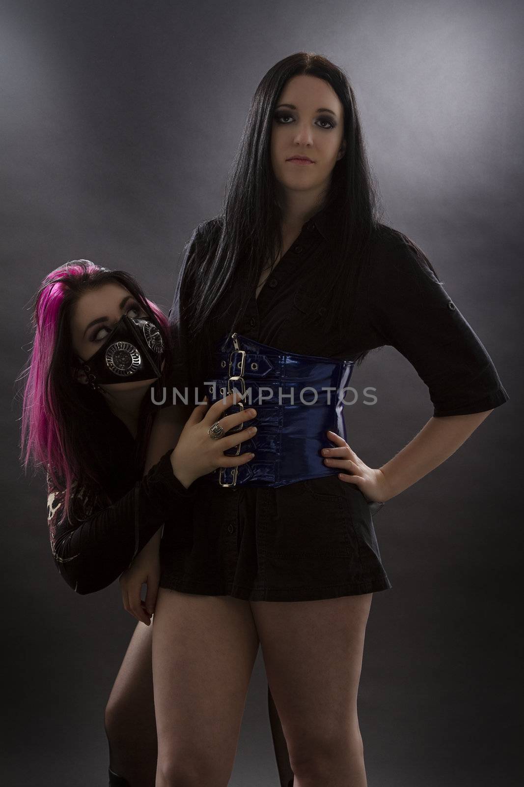 crazy looking teenage girl wearing goth inspired clothes with pink and black hair and gas mask, being shy and hidding behind her mistress