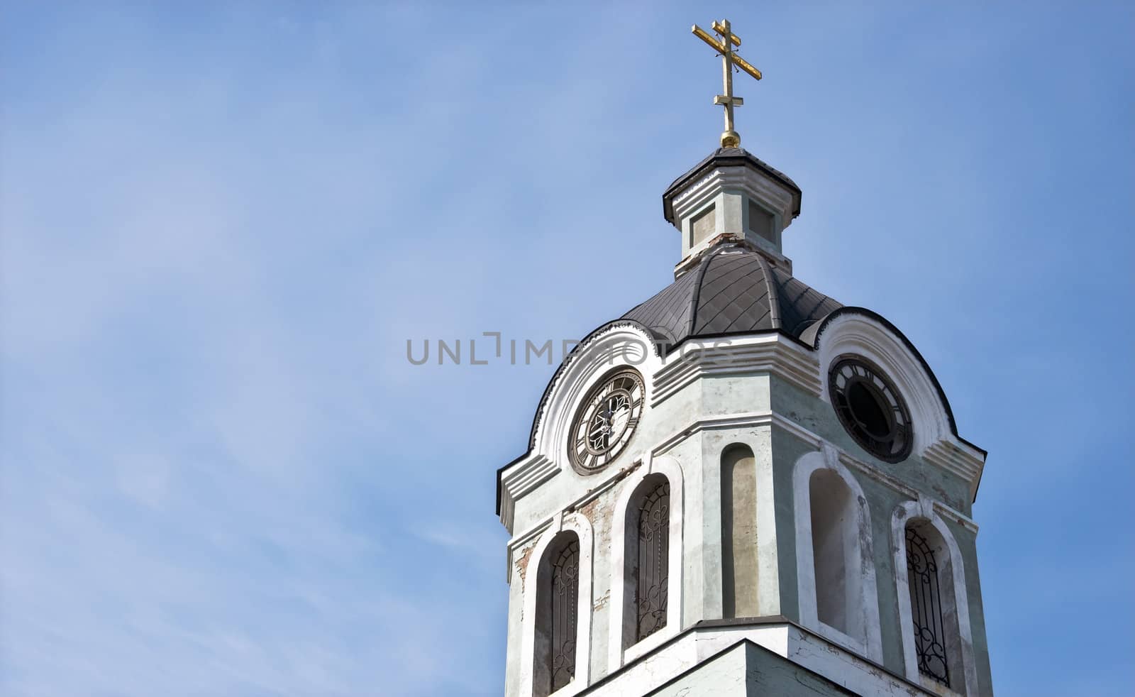 Church bell tower with a clock and a cross against the blue sky.