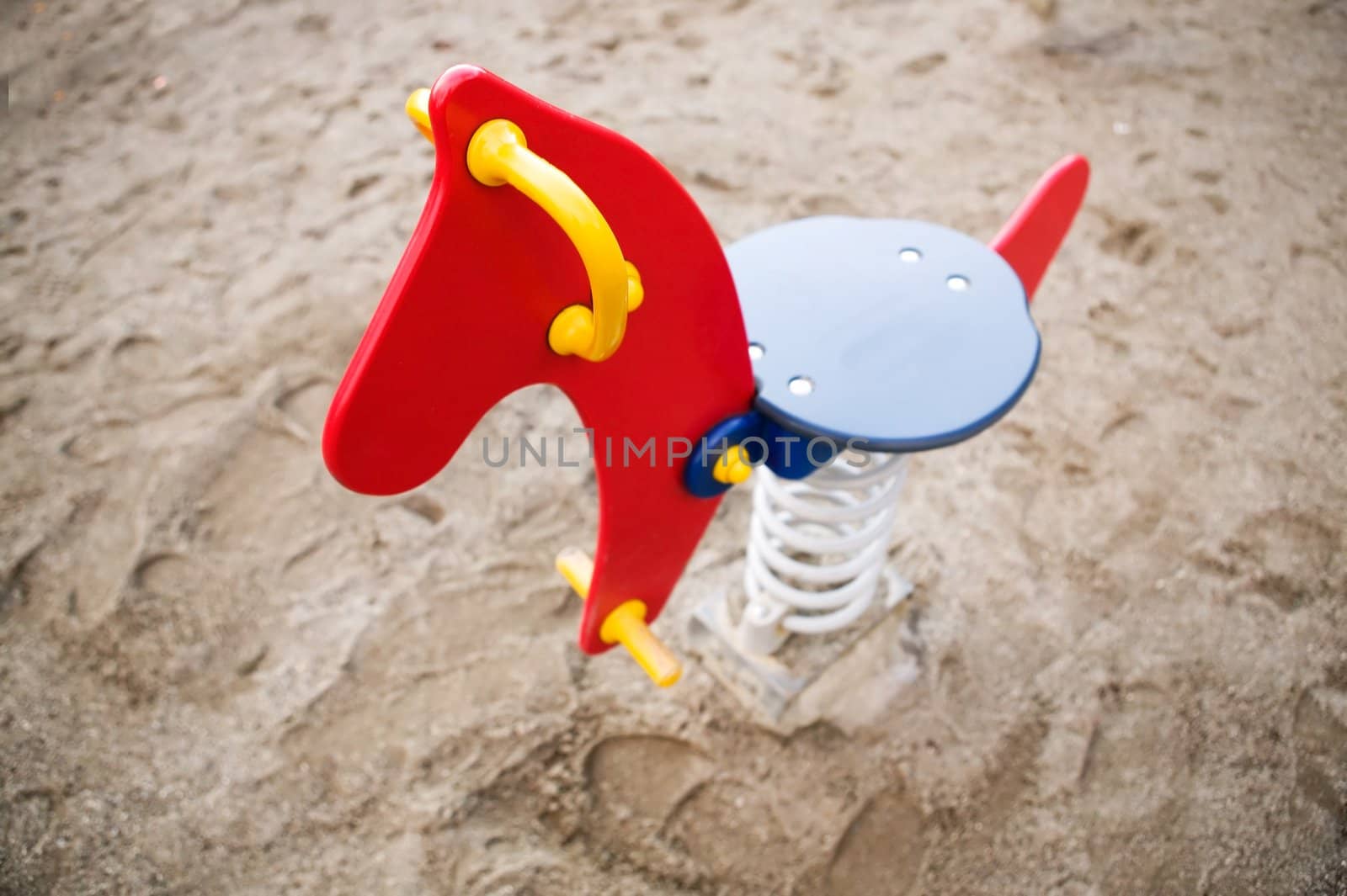 A brightly colored red, blue and yellow rocking horse in a public playground surrounded by sand. - focus on the head with a shallow depth of field.