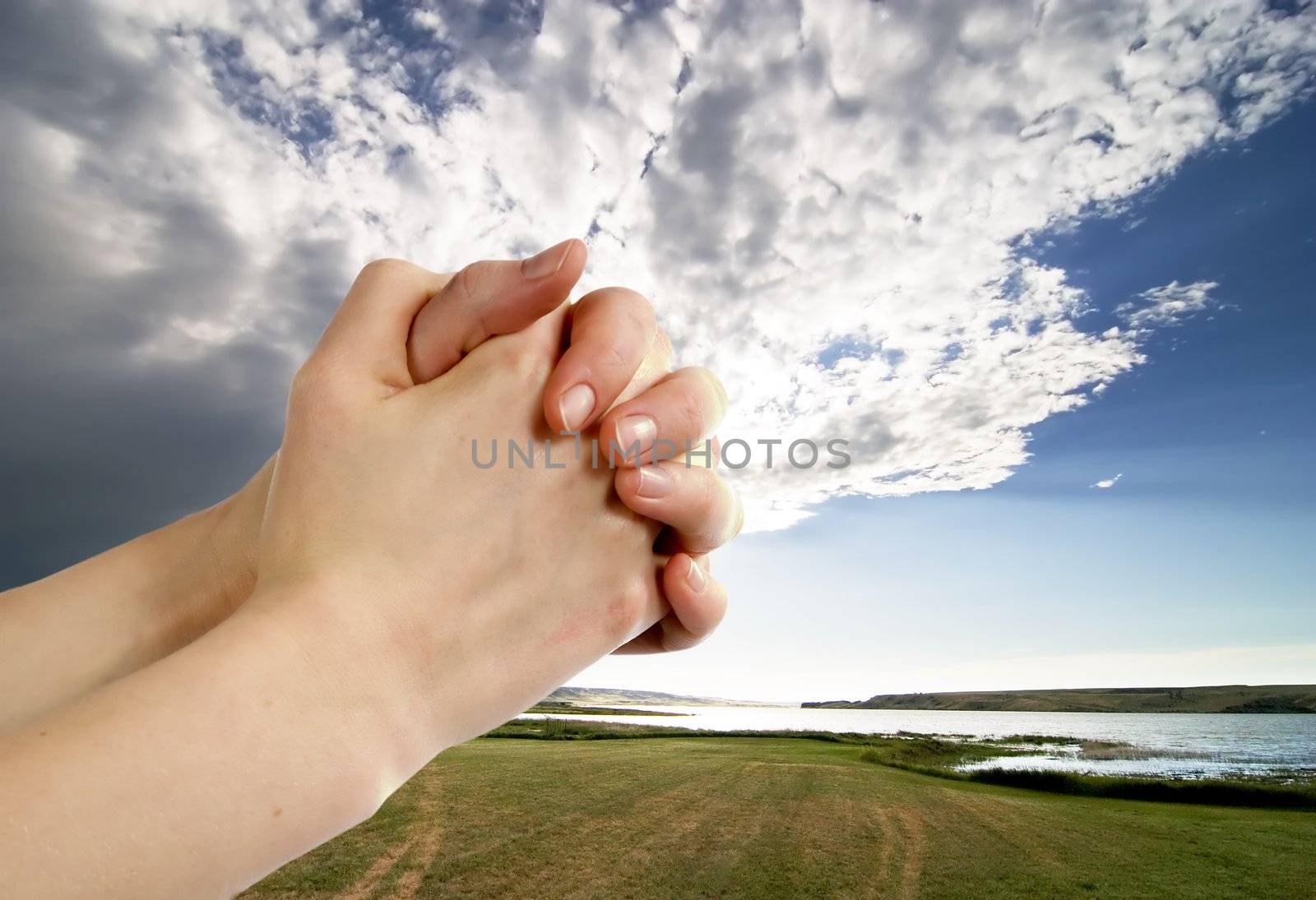 A pair of hands in a praying position set against a beautiful prairie lake landscape