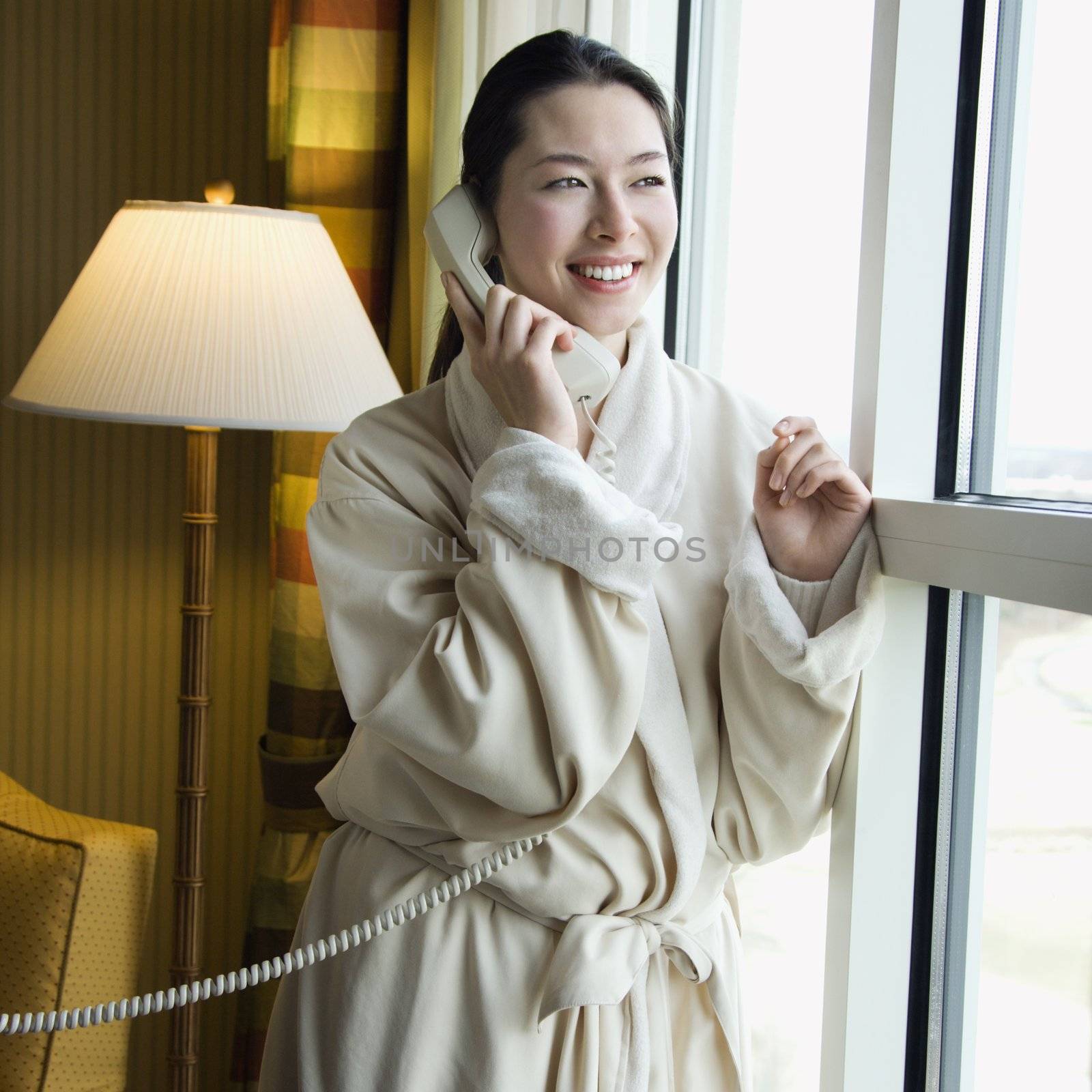 Taiwanese mid adult woman in bathrobe talking on phone and looking out window.