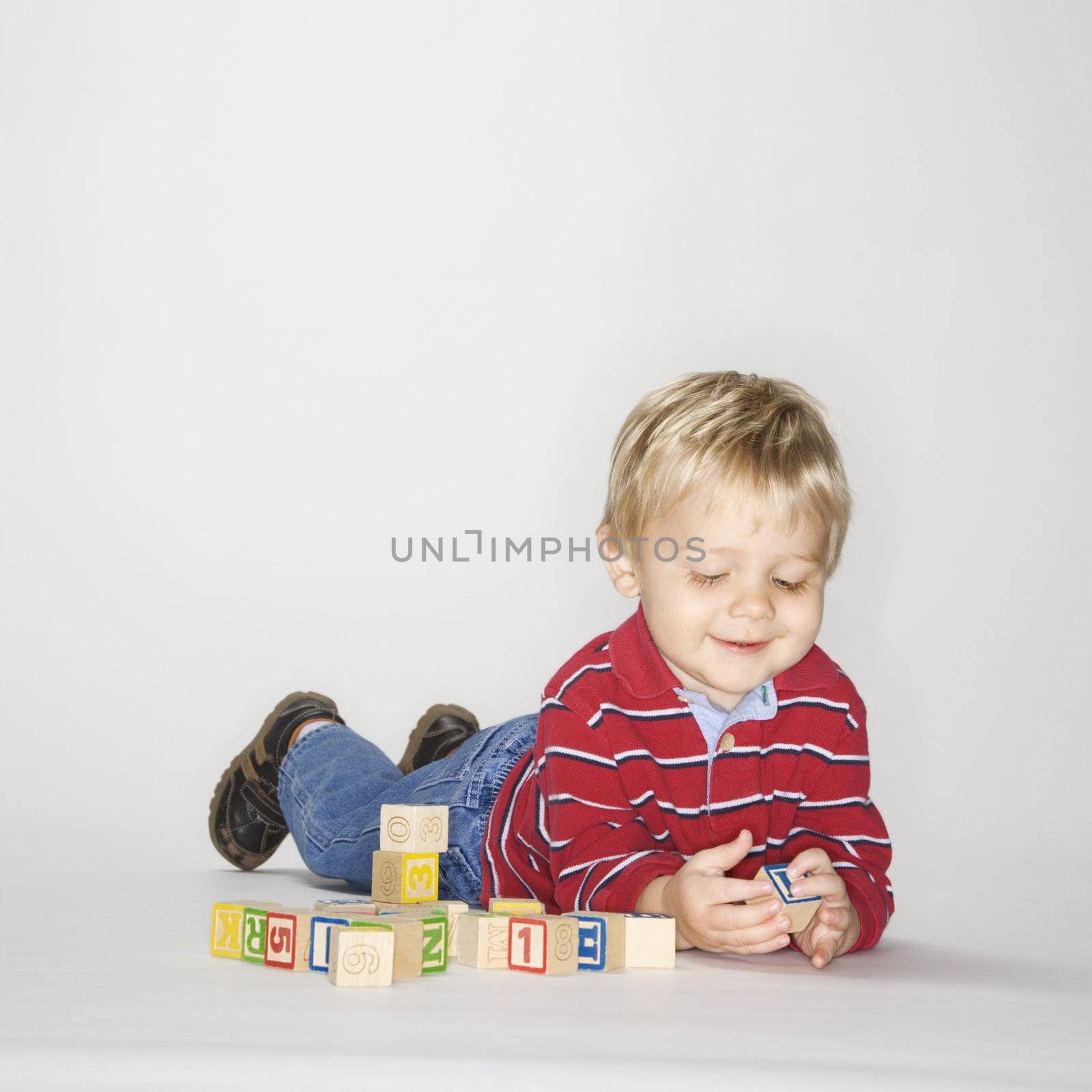 Studio portrait of Caucasian boy lying on stomach propped up on elbows playing with toy blocks.