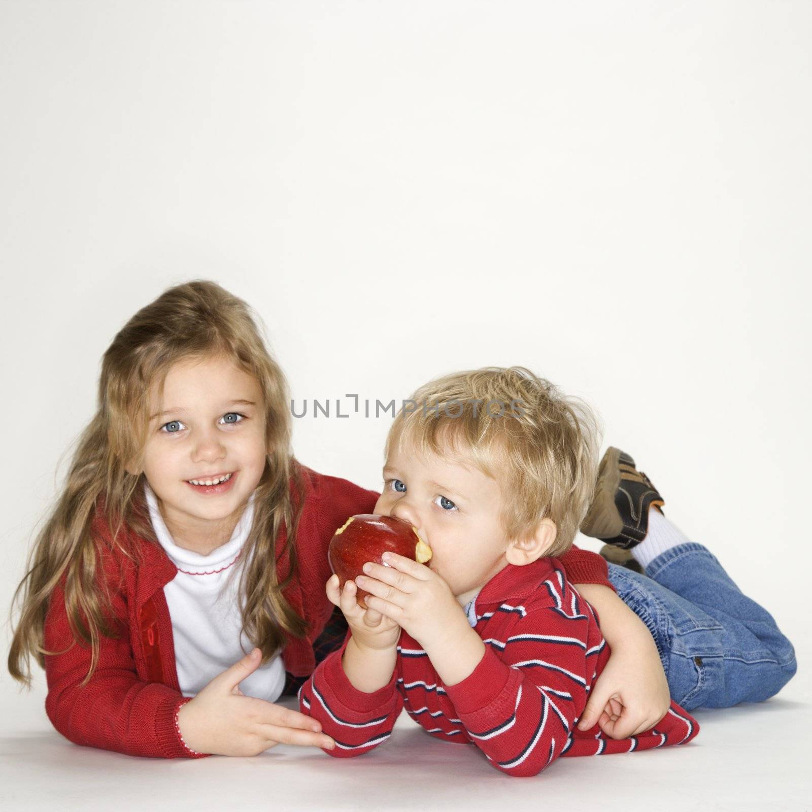 Studio portrait of Caucasian girl with arm around boy eating apple against white background.