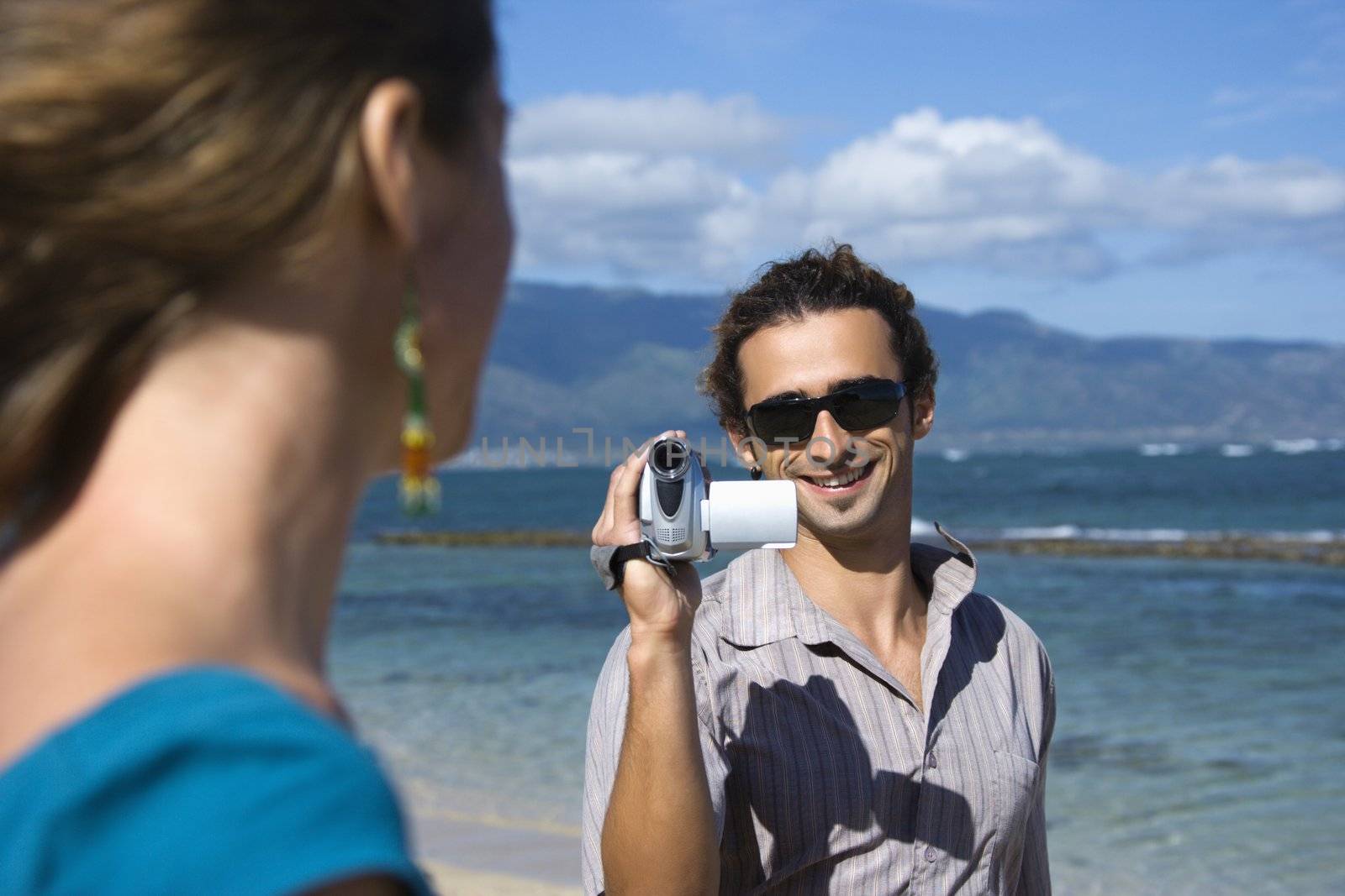 Mid-adult Caucasian man on beach pointing video camera at woman.