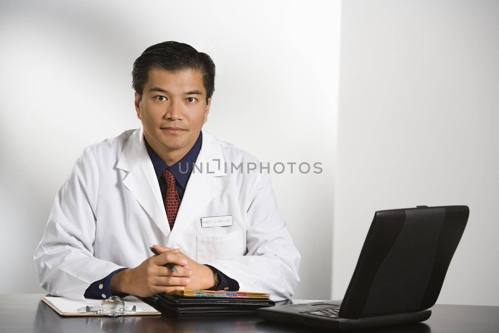 Asian American male doctor sitting at desk with charts and laptop computer looking at viewer.