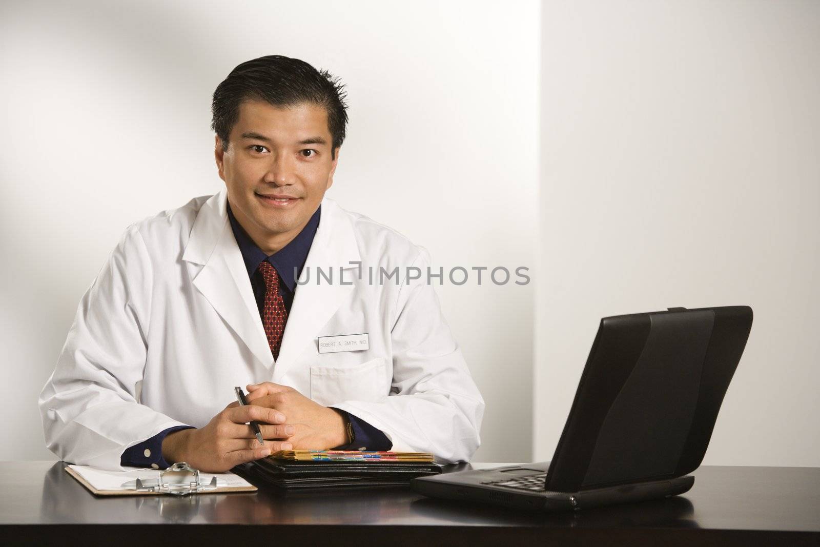 Asian American male doctor sitting at desk with charts and laptop computer looking at viewer.