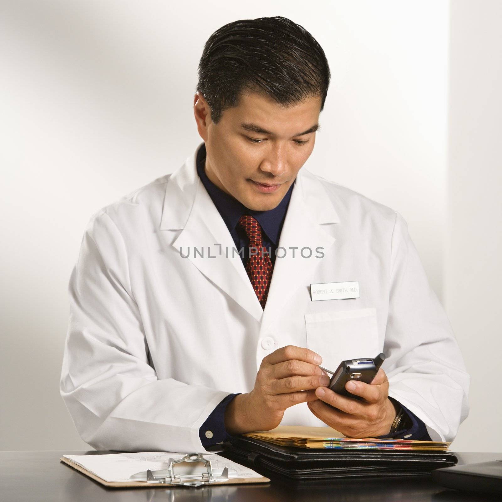 Asian American male doctor sitting at desk with charts using pda.