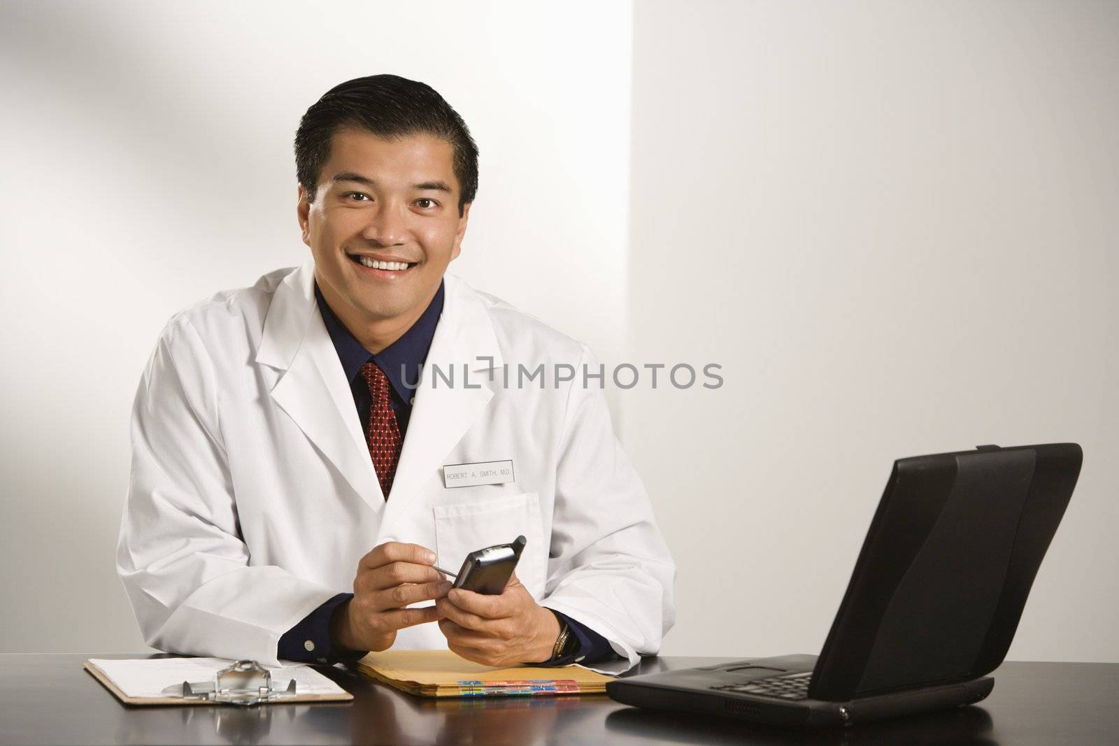 Asian American male doctor sitting at desk with charts and laptop computer using pda.