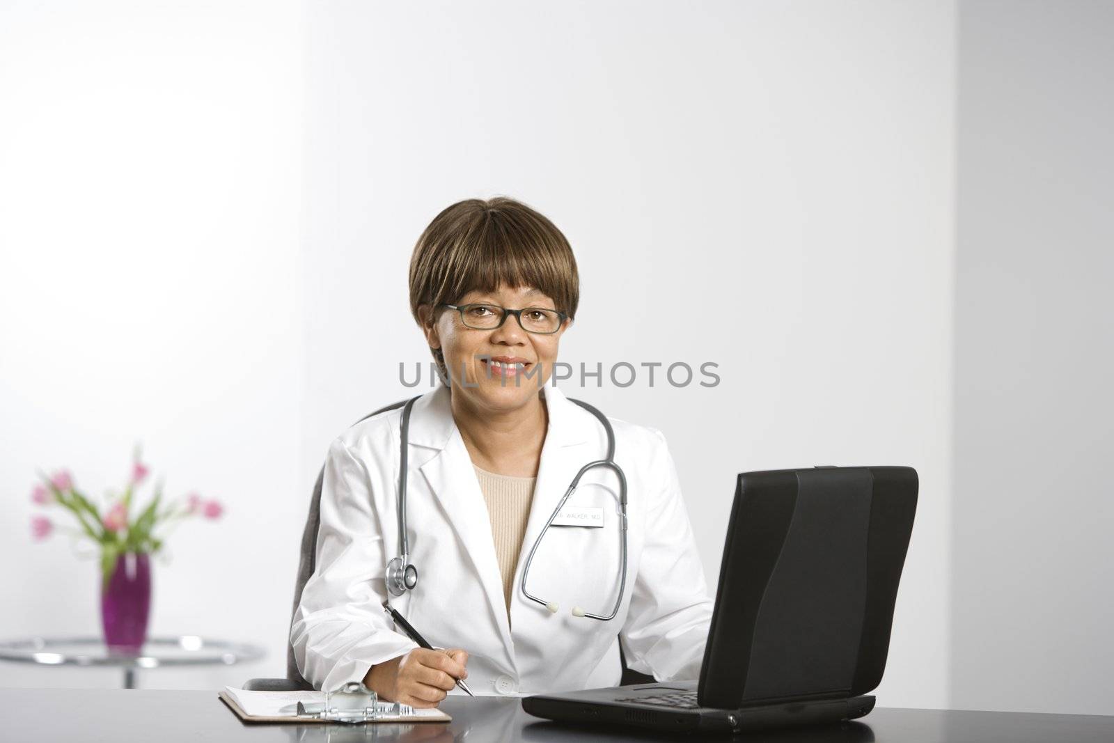 African American middle-aged female doctor sitting at desk working on laptop, smiling and looking at viewer.