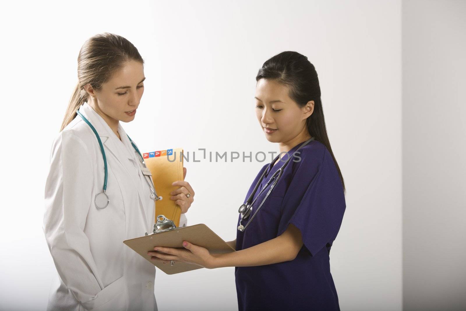 Caucasian mid-adult female doctor and Asian Chinese mid-adult female physician's assistant discussing paperwork.