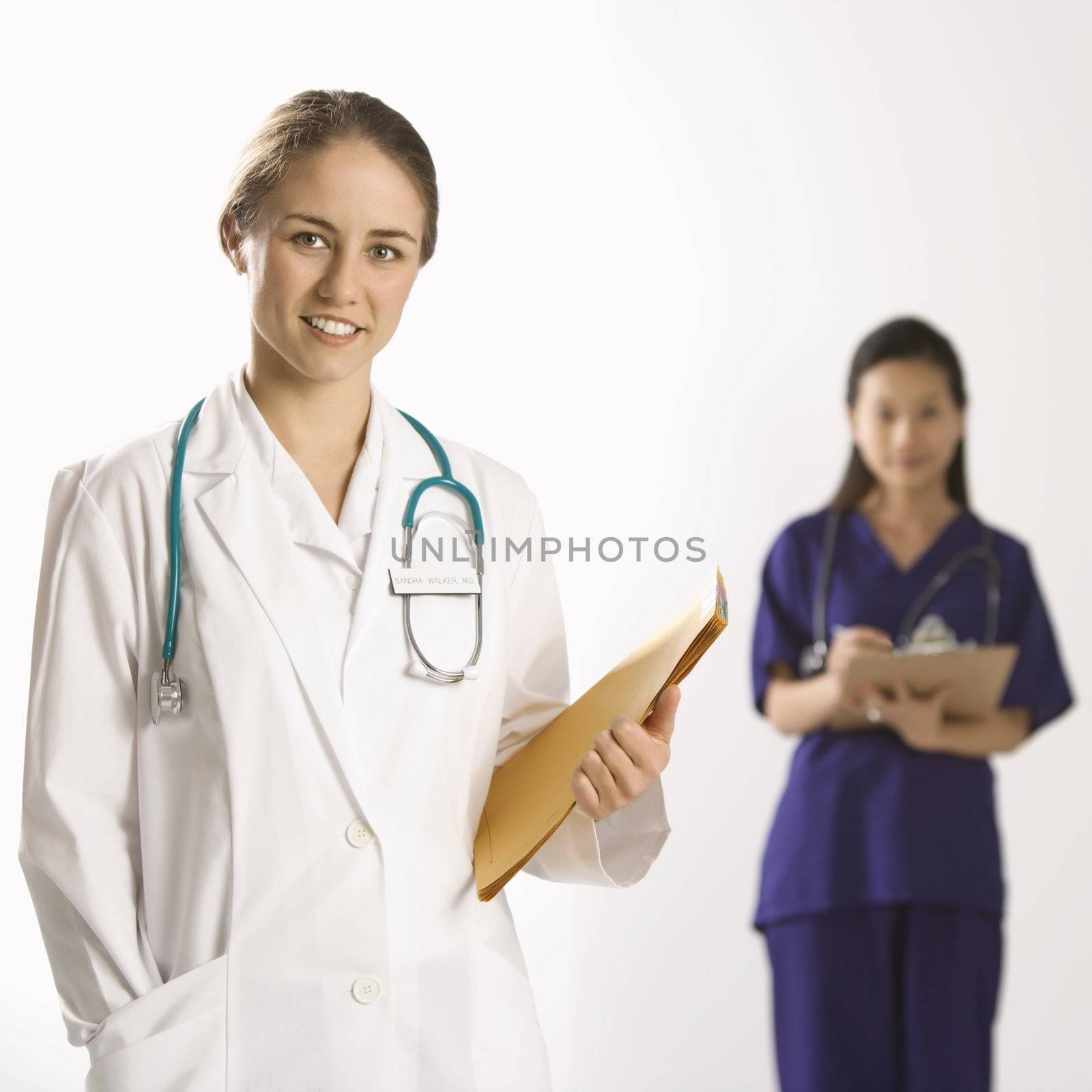 Caucasian mid-adult female doctor smiling and looking at viewer with Asian Chinese mid-adult female physician's assistant standing in background.