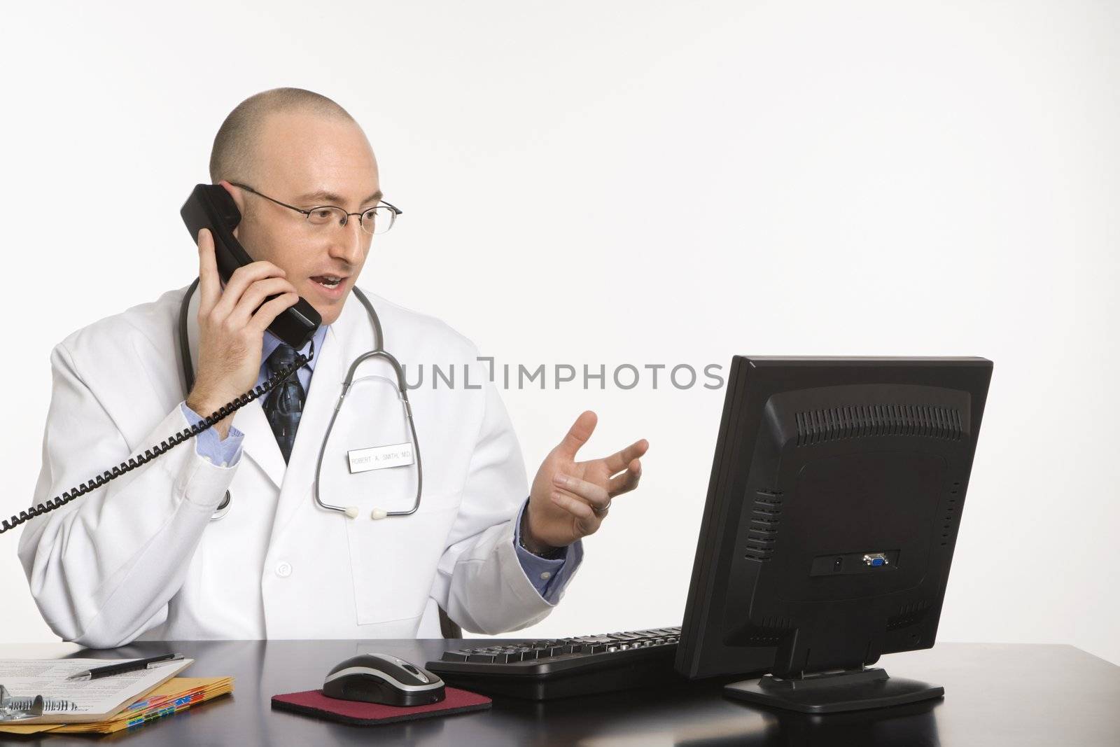 Caucasian mid adult male physician sitting at desk with laptop computer talking on telephone.