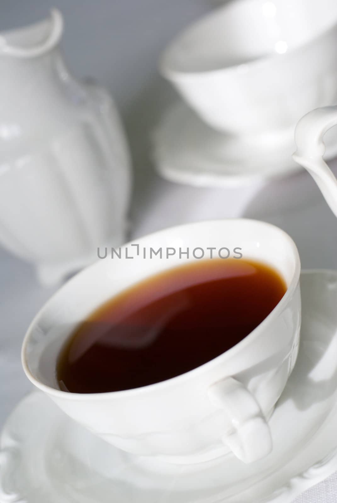 Cup of tea (focus only in the middle part of the cup edge) and porcelain milk jug and another cup behind on the white tablecloth. Shallow depth of field.